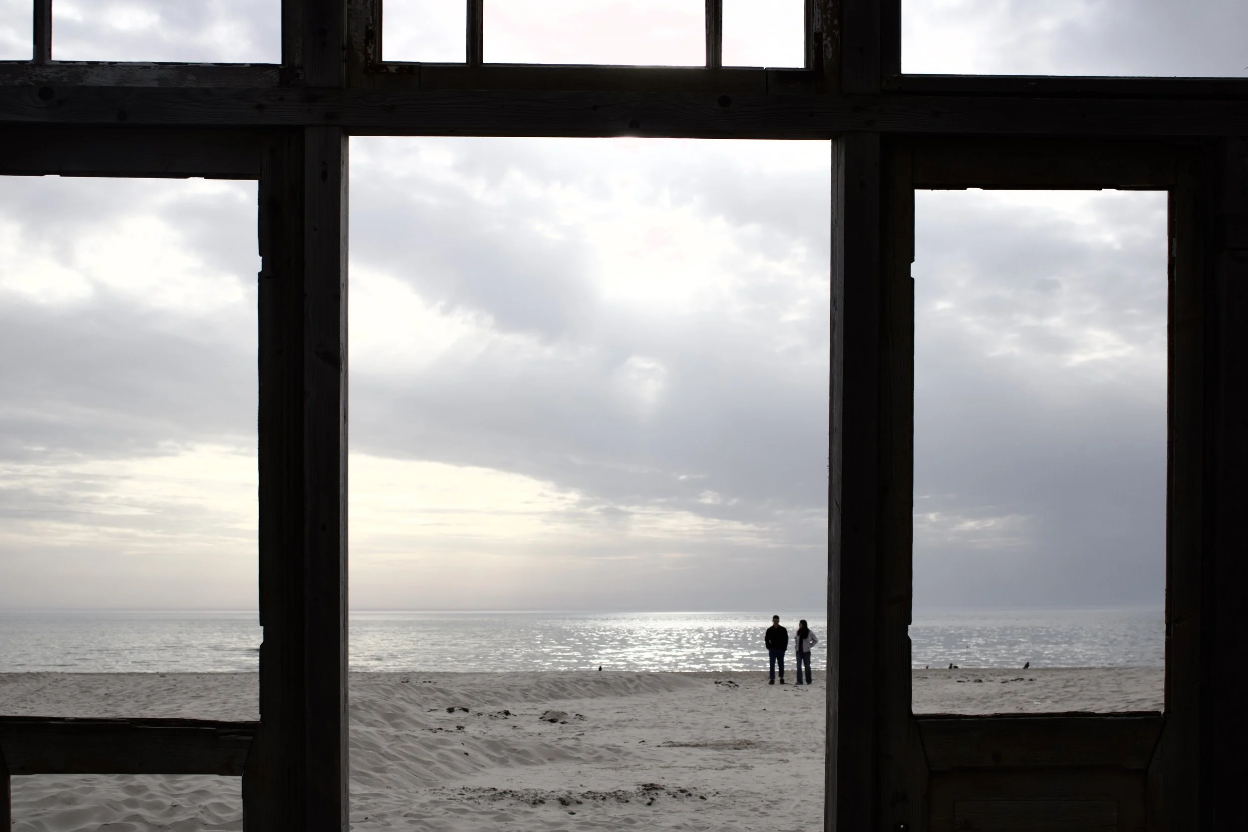 View through a wooden frame at the beach showing two people standing by the shoreline with overcast sky and ocean in the background.