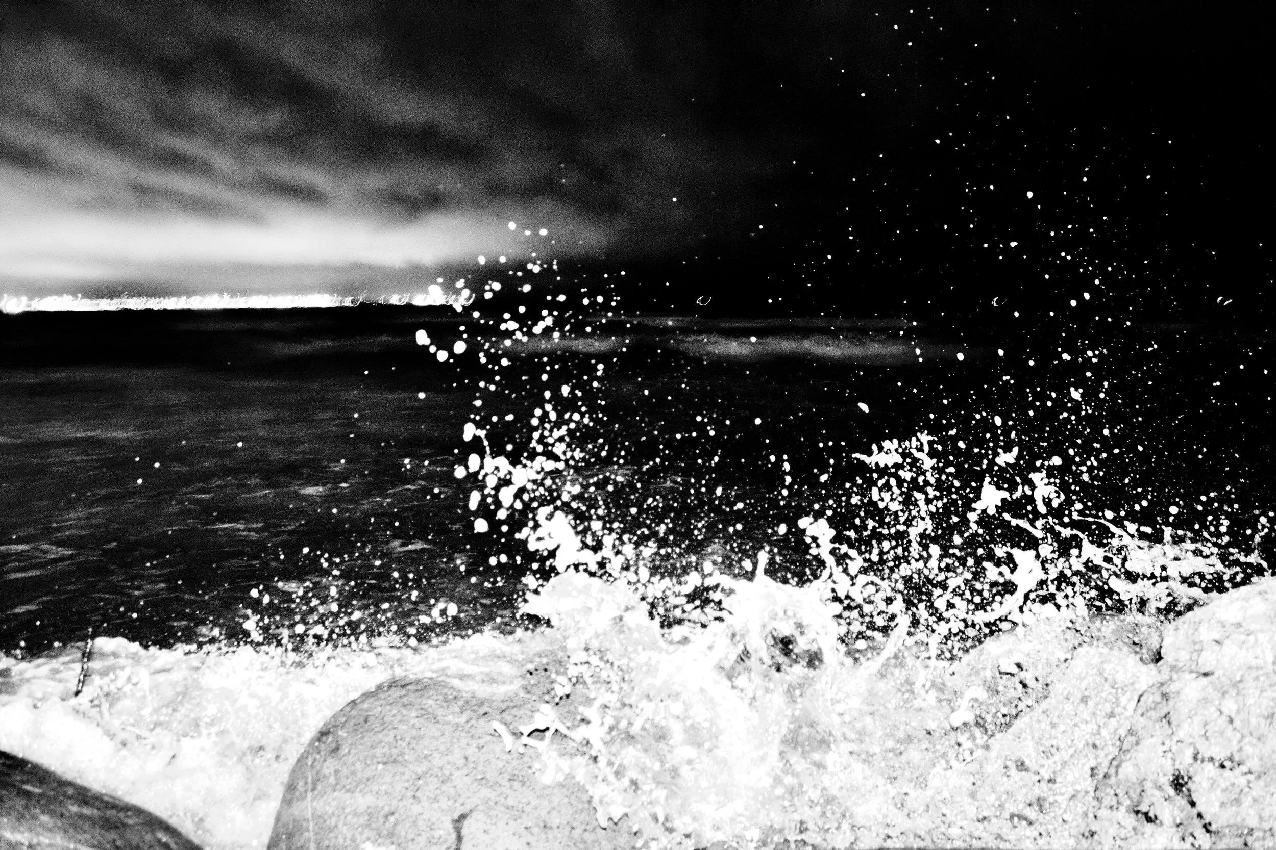 Photo of ocean waves crashing on the sandy shore at night, with spray and foam in the air and dark sky above.