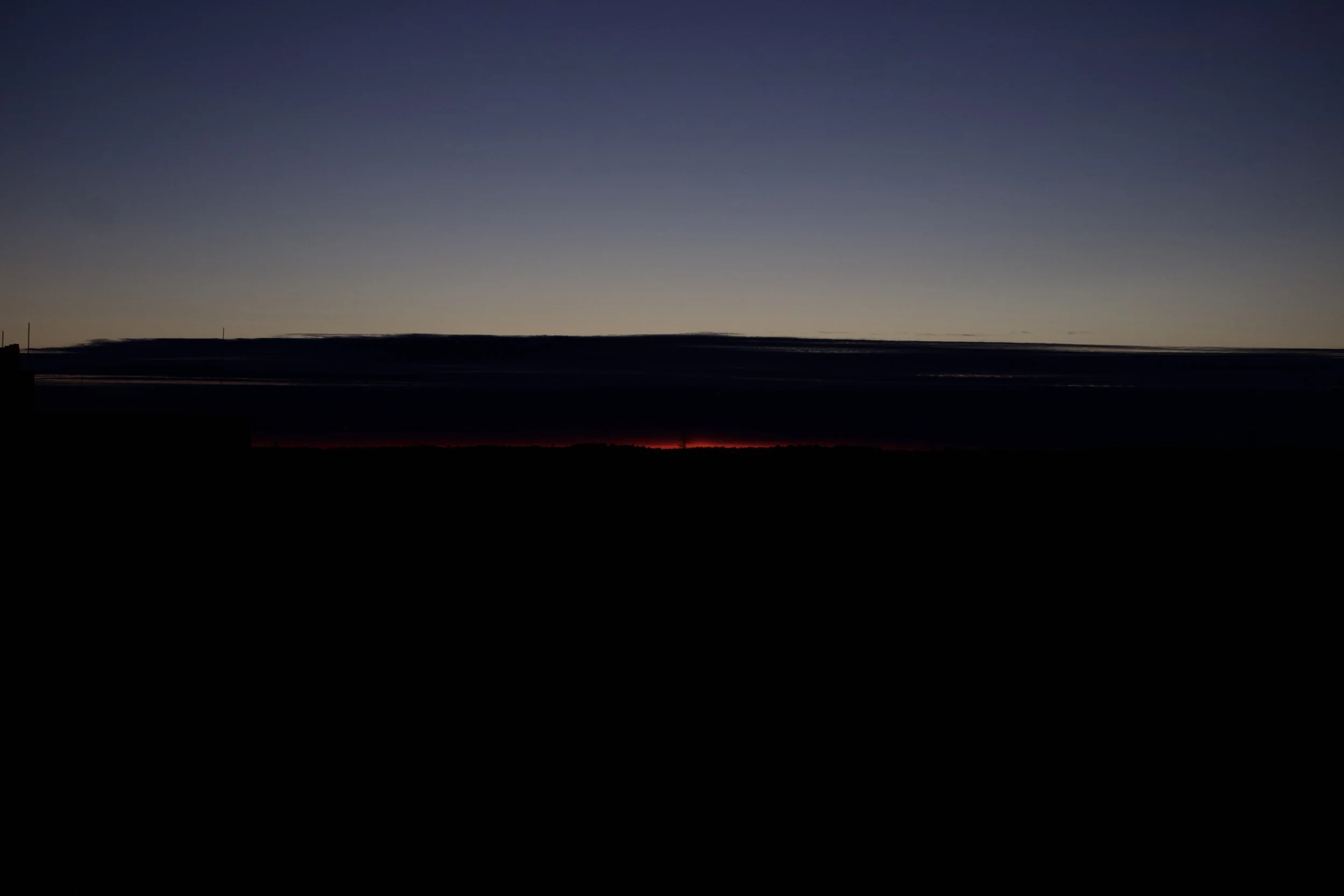 A dark landscape with a horizon glow at sunset or sunrise, featuring a mostly clear sky with subtle gradient colors.