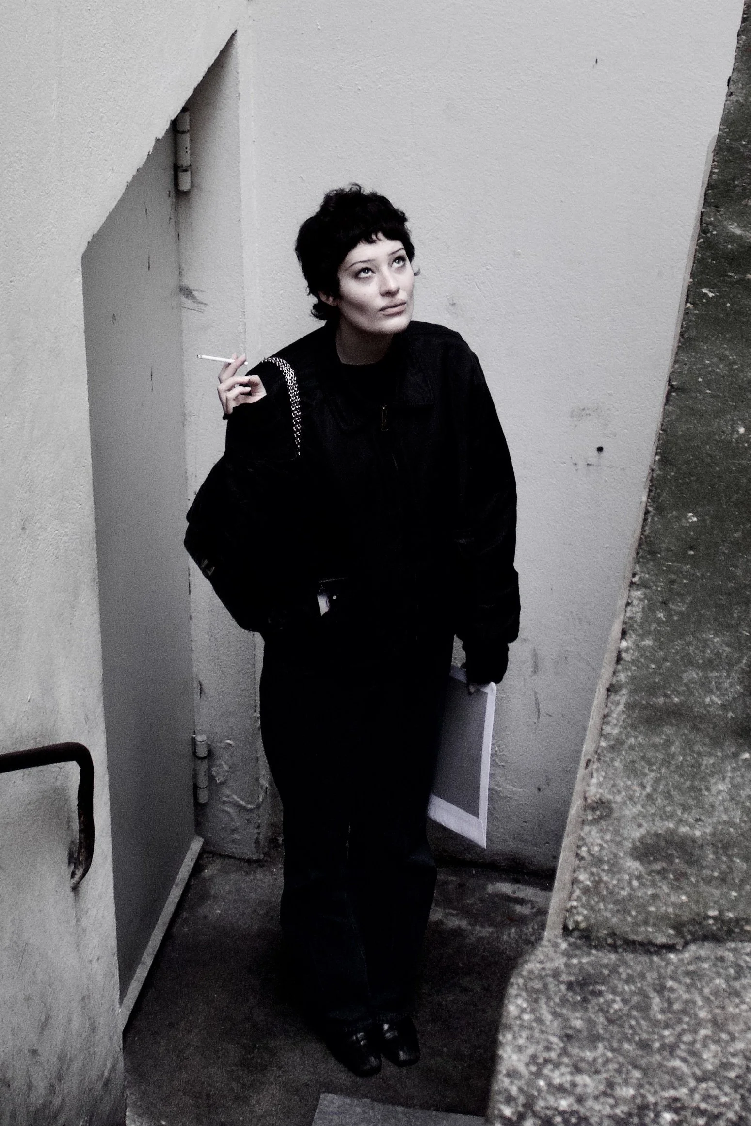 A woman with short dark hair holding a cigarette, standing in a corner between a greenish wall and a plain off-white wall, wearing a black jacket and black pants.