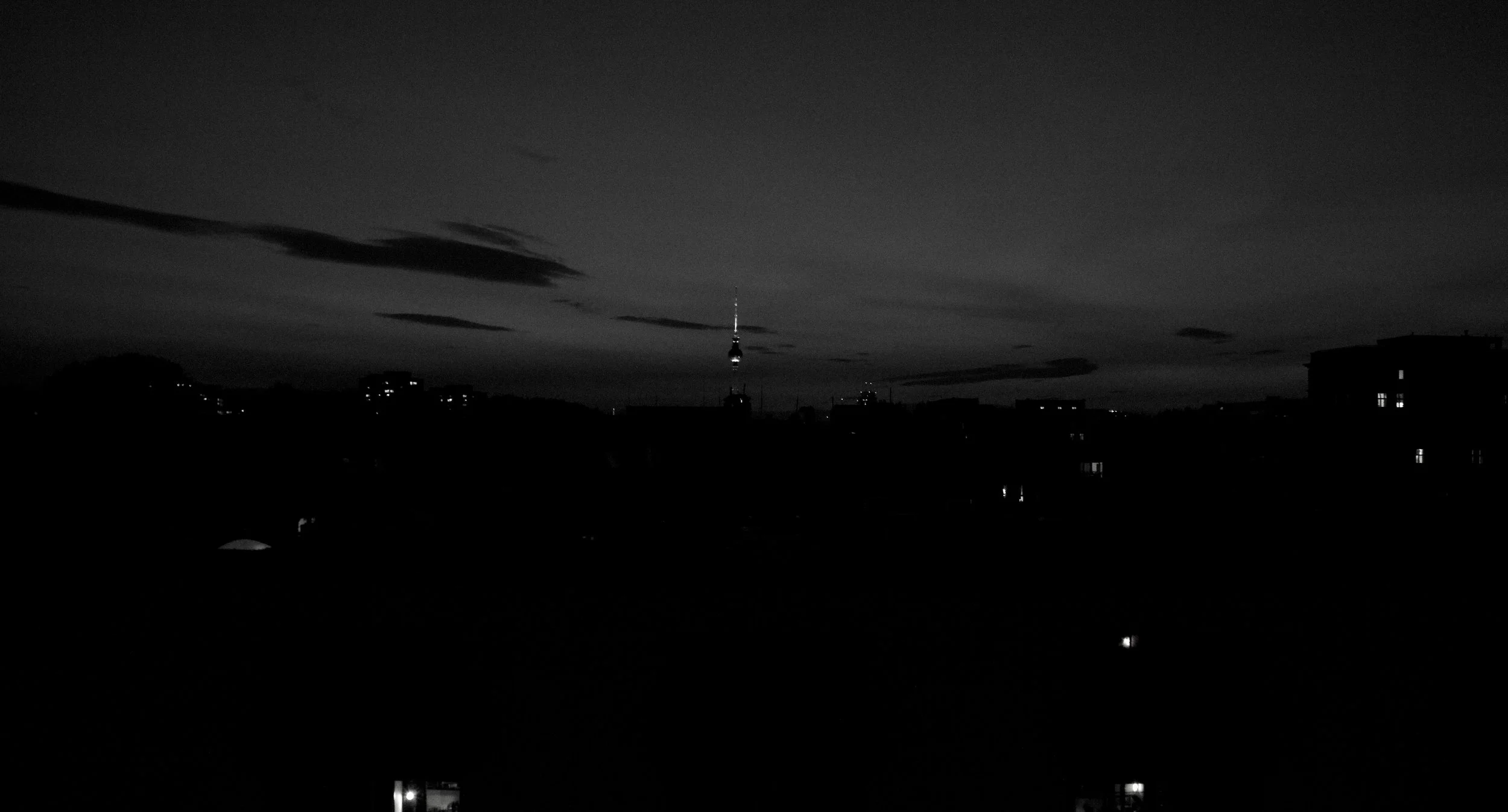 Nighttime cityscape in black and white, with a television tower in the distance and a cloudy sky.