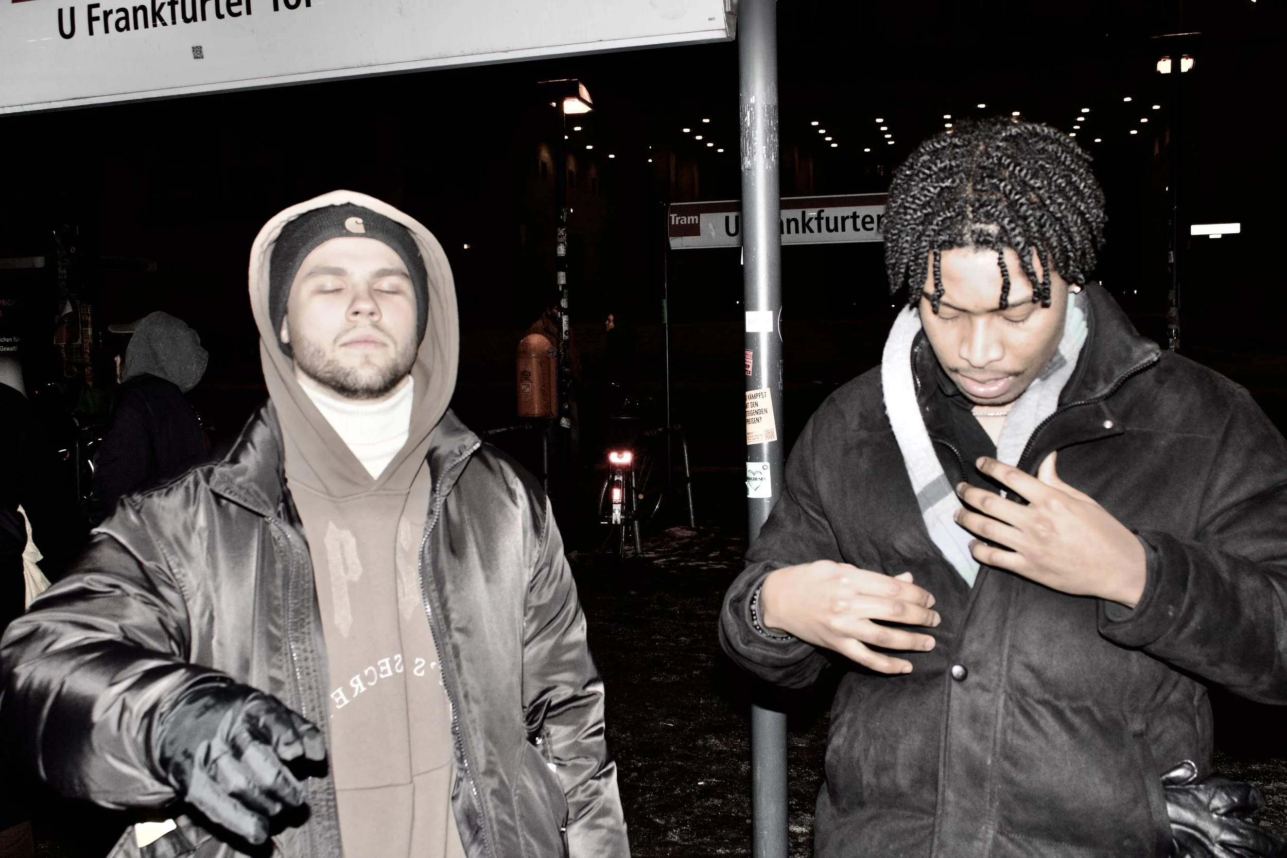 Two young men standing at night in front of a metro station sign that reads 'U Frankfurt' and a street sign that says 'U Frankfurter.' One man, on the left, has his eyes closed, wearing a hoodie under a jacket, and the other man, on the right, has cu