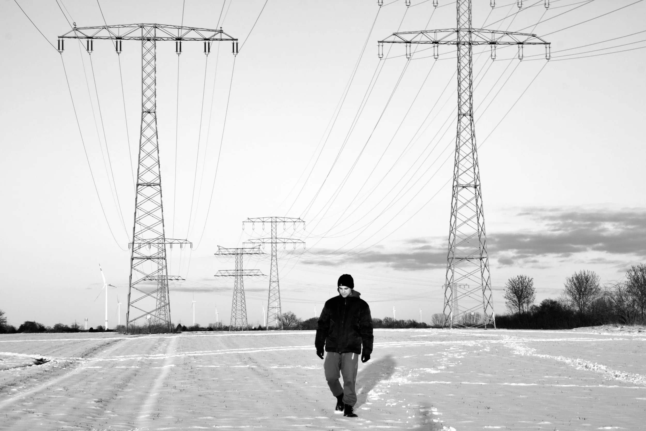 A person walking on an open area with power lines and wind turbines in the background.