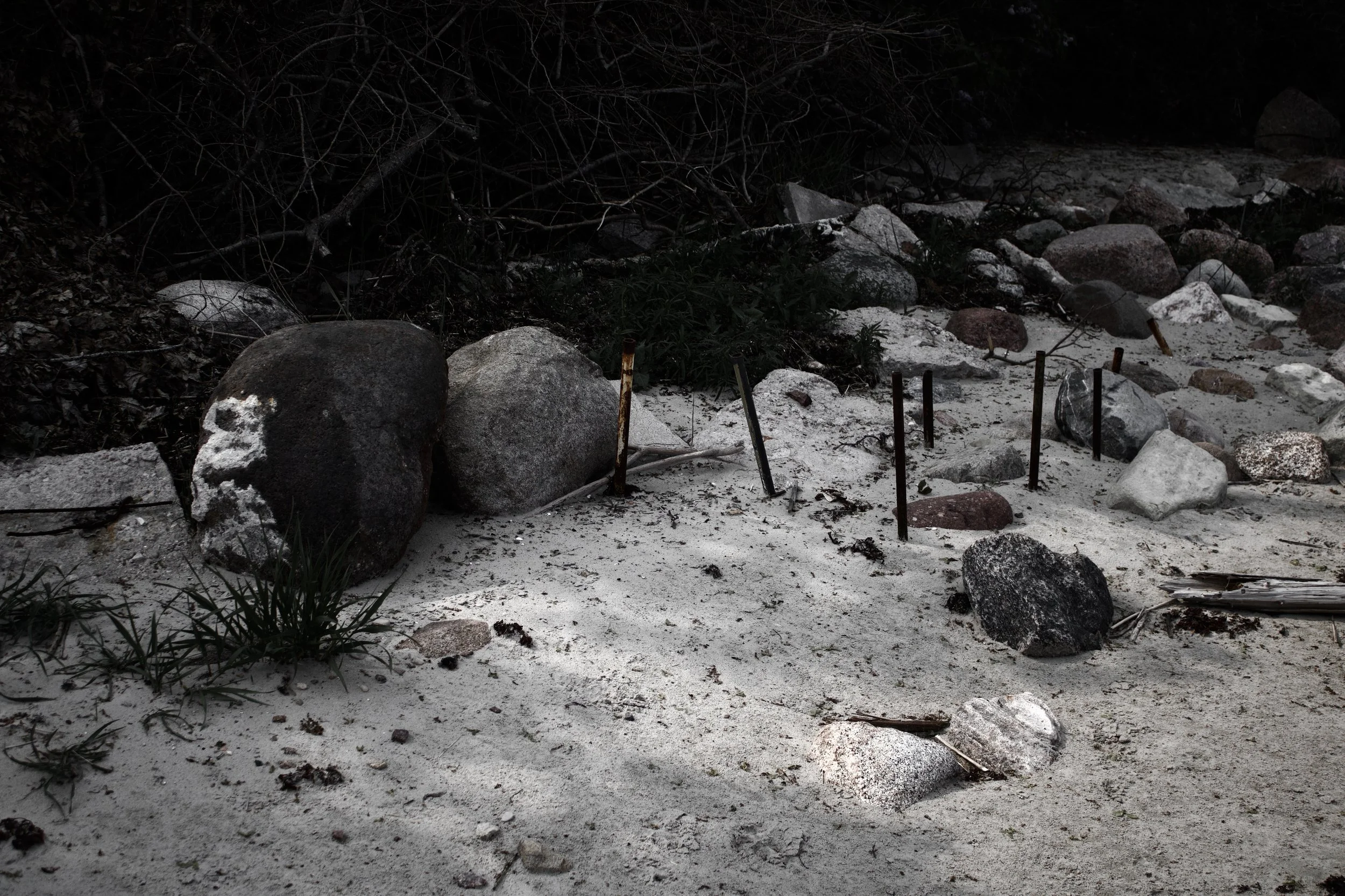 A rocky beach with various large stones and smaller rocks, some rusted metal rods sticking out of the sand, and bushes and dark trees in the background.