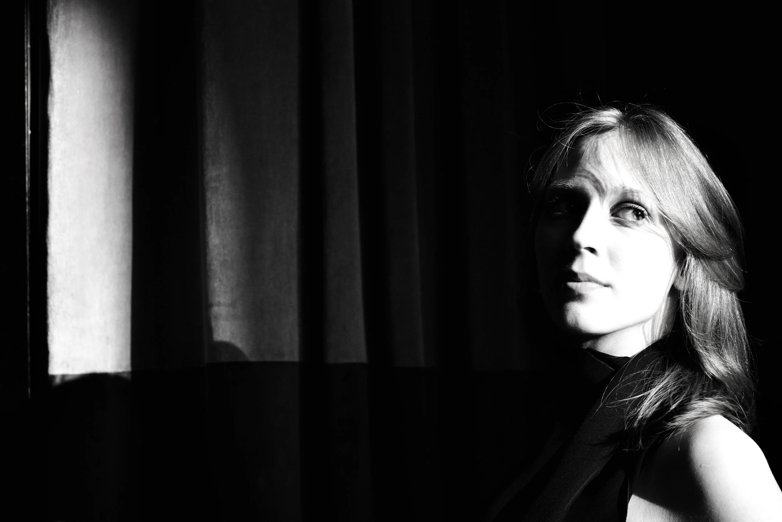 Black and white photo of a young girl with long hair, looking out a window with blinds, partially illuminated, with shadows on her face.