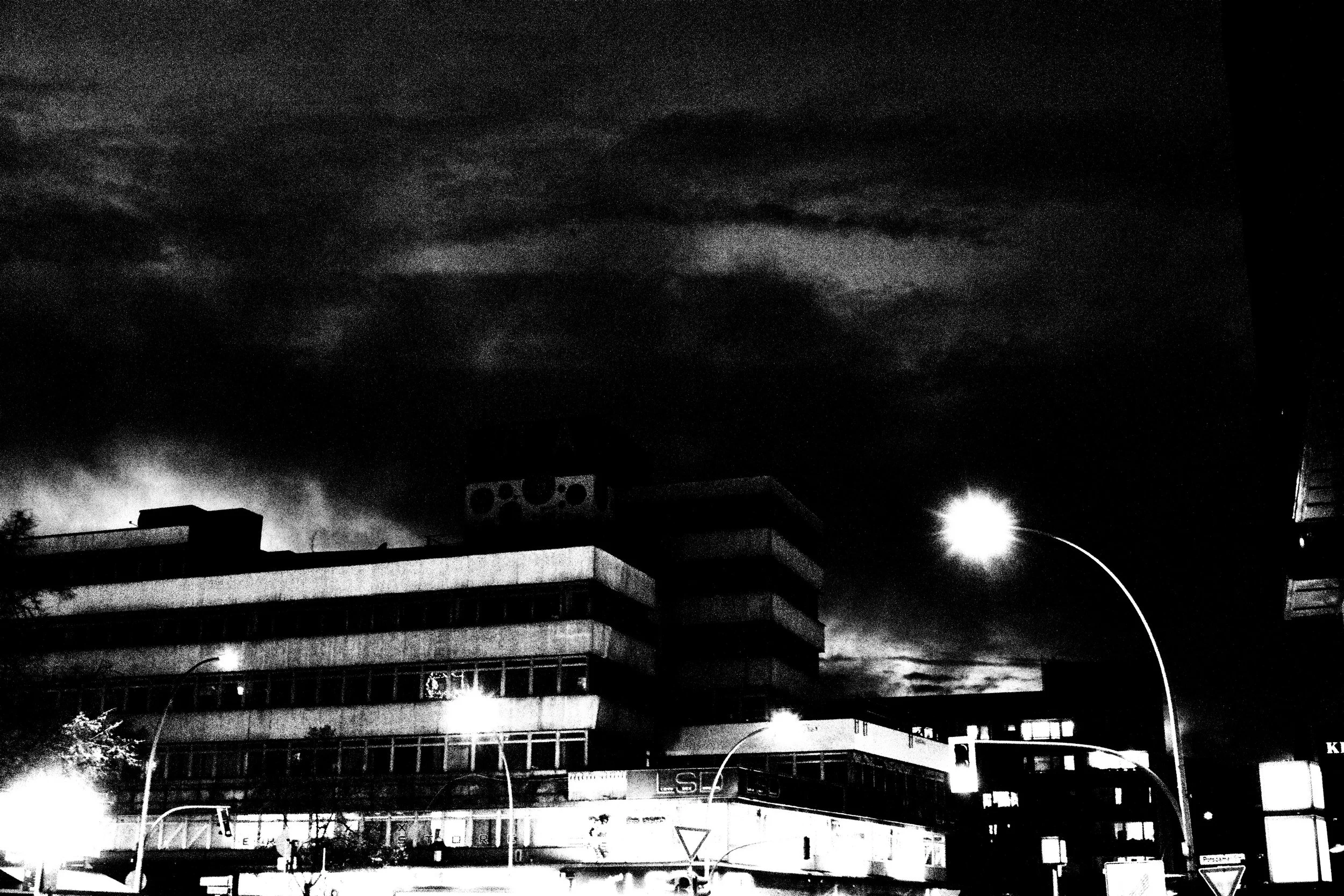 Black and white photo of a cityscape at night with buildings, streetlights, and a dark cloudy sky.