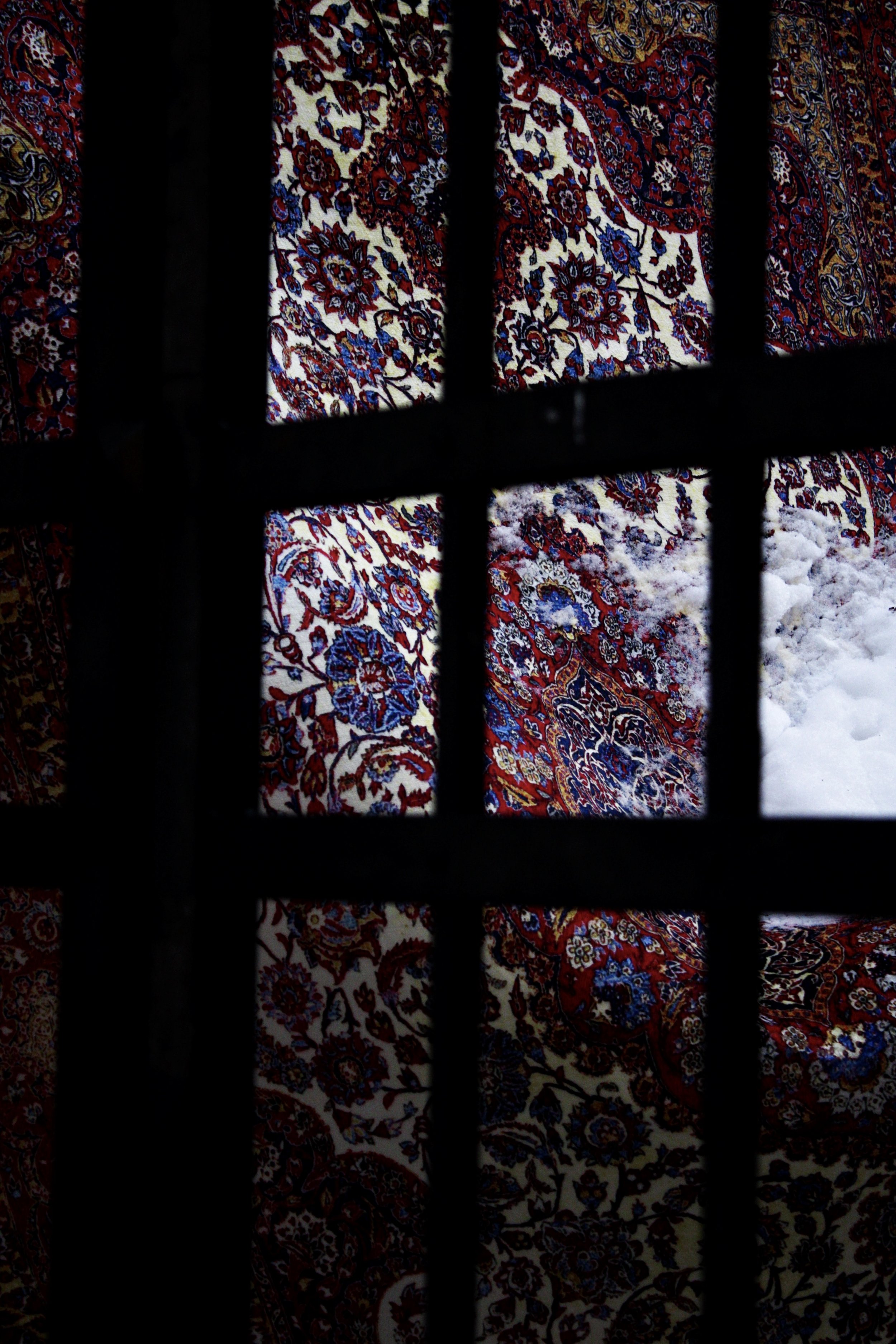 A hanging carpet with floral designs in rich colours. It's seen through a grid-like black frame.
