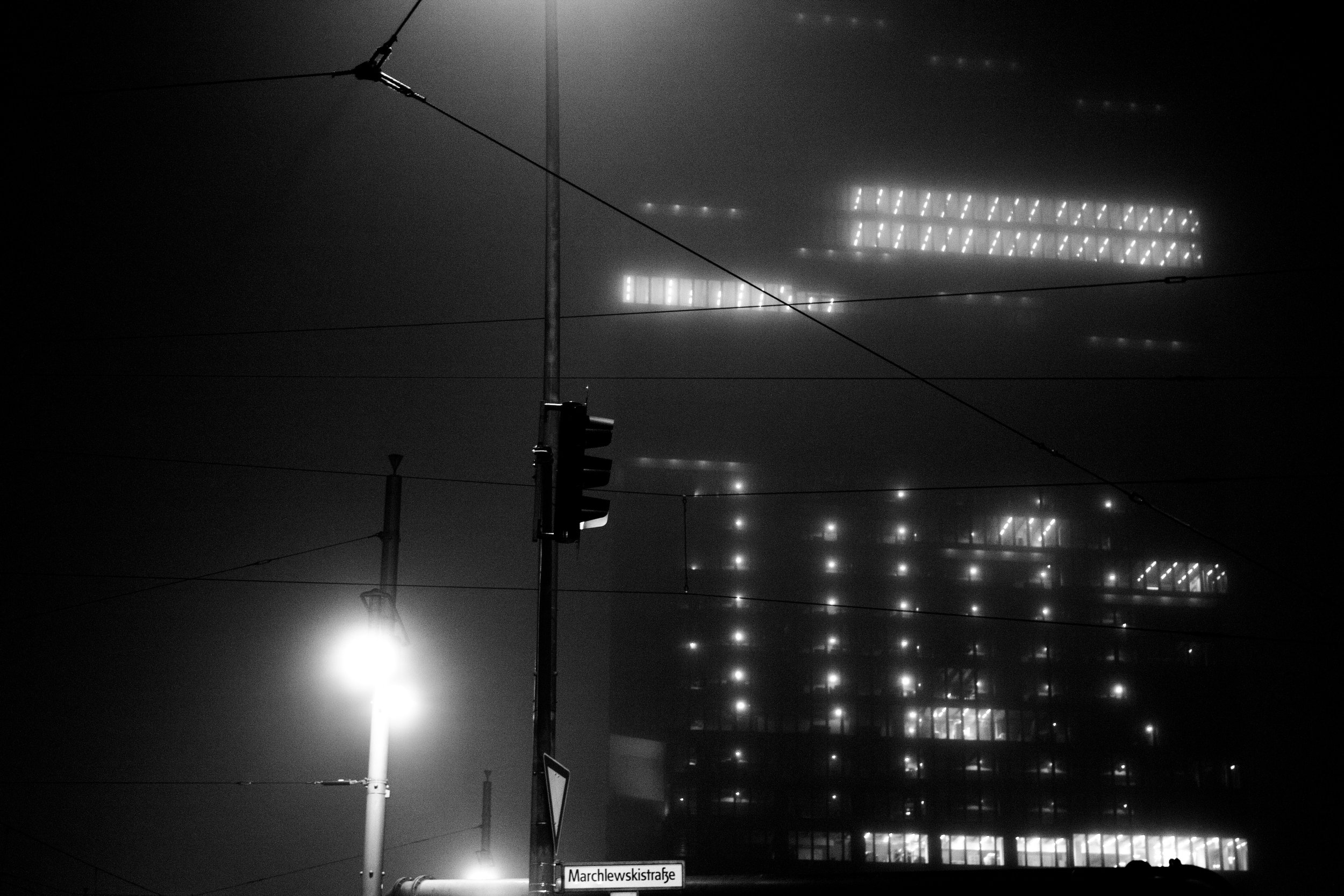 Black and white photo of a foggy city street with traffic lights, a street sign reading 'Marchlewski strasse,' and a modern building with illuminated windows in the background.