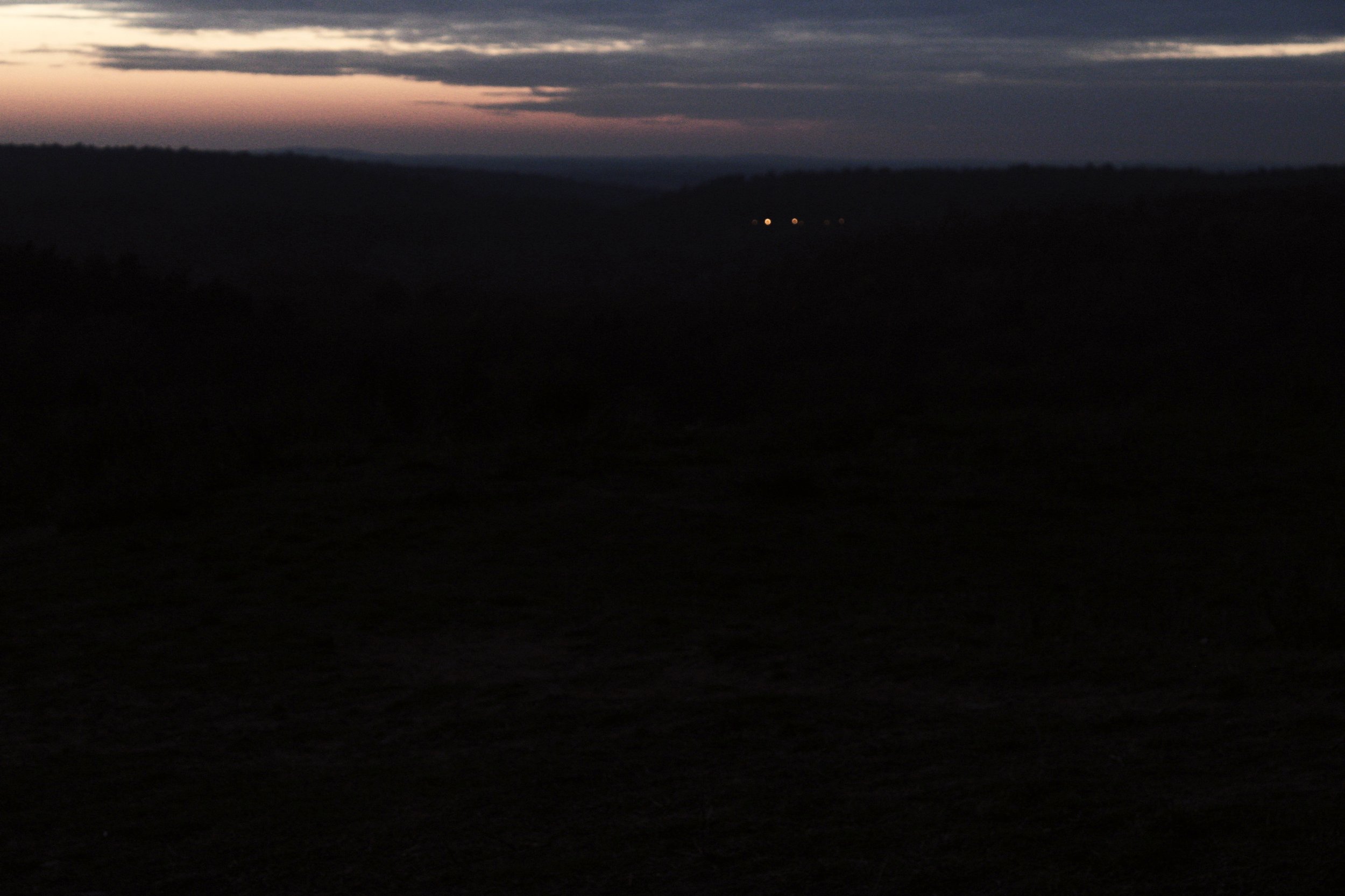 Hilly landscape shortly after the sunset, with several feeble lights shining in the distance.