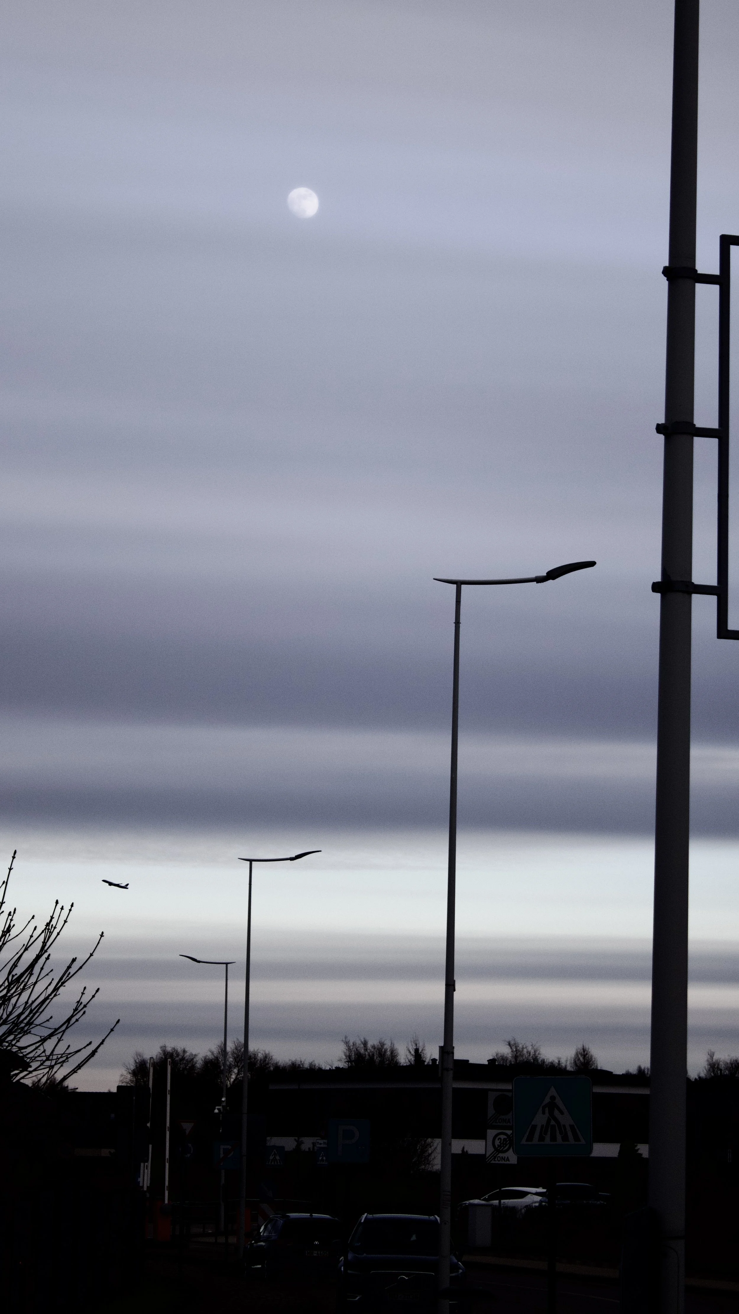 Overcast sky with the moon visible, street lamps, and a few airplanes in the distance, with cars parked in a parking lot at night.
