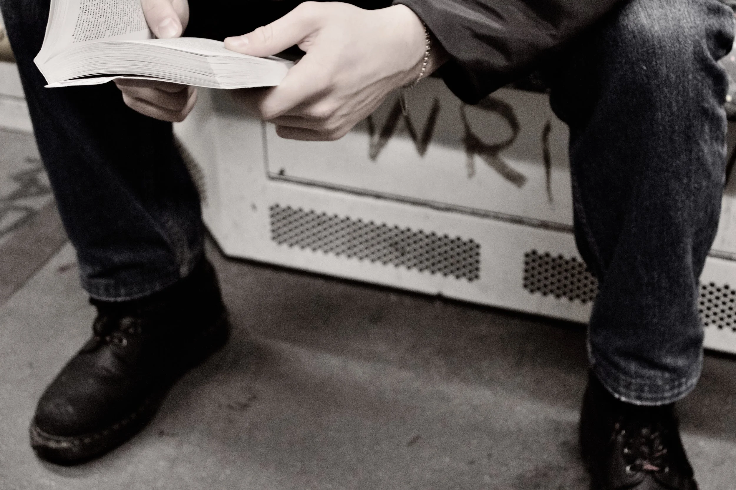 Person sitting on a bench reading a book, with their legs apart wearing black boots and dark jeans, and holding the book with both hands.