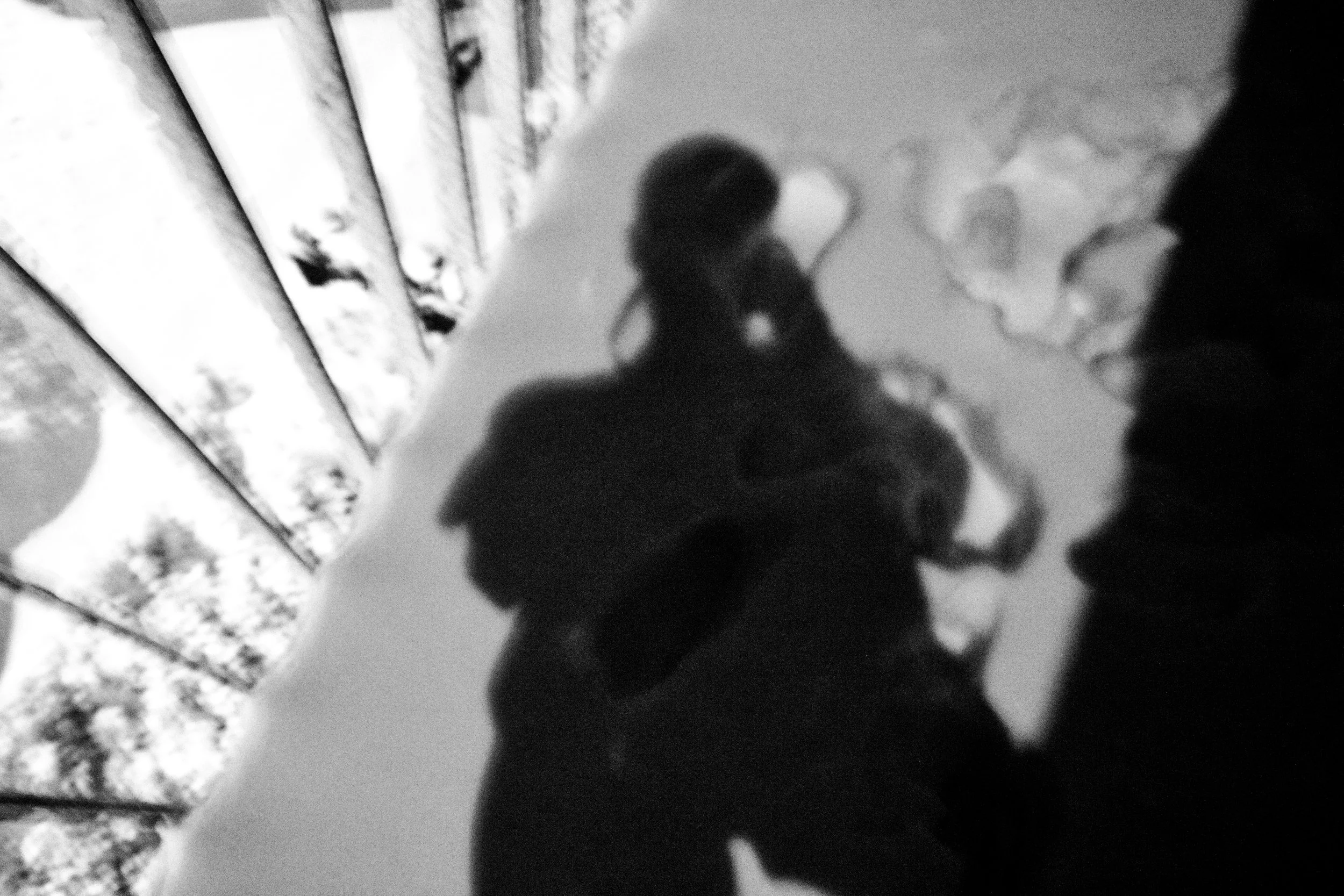 Black and white photo of a person taking a selfie, with a patterned curtain on the left and a background with soft, out-of-focus elements.