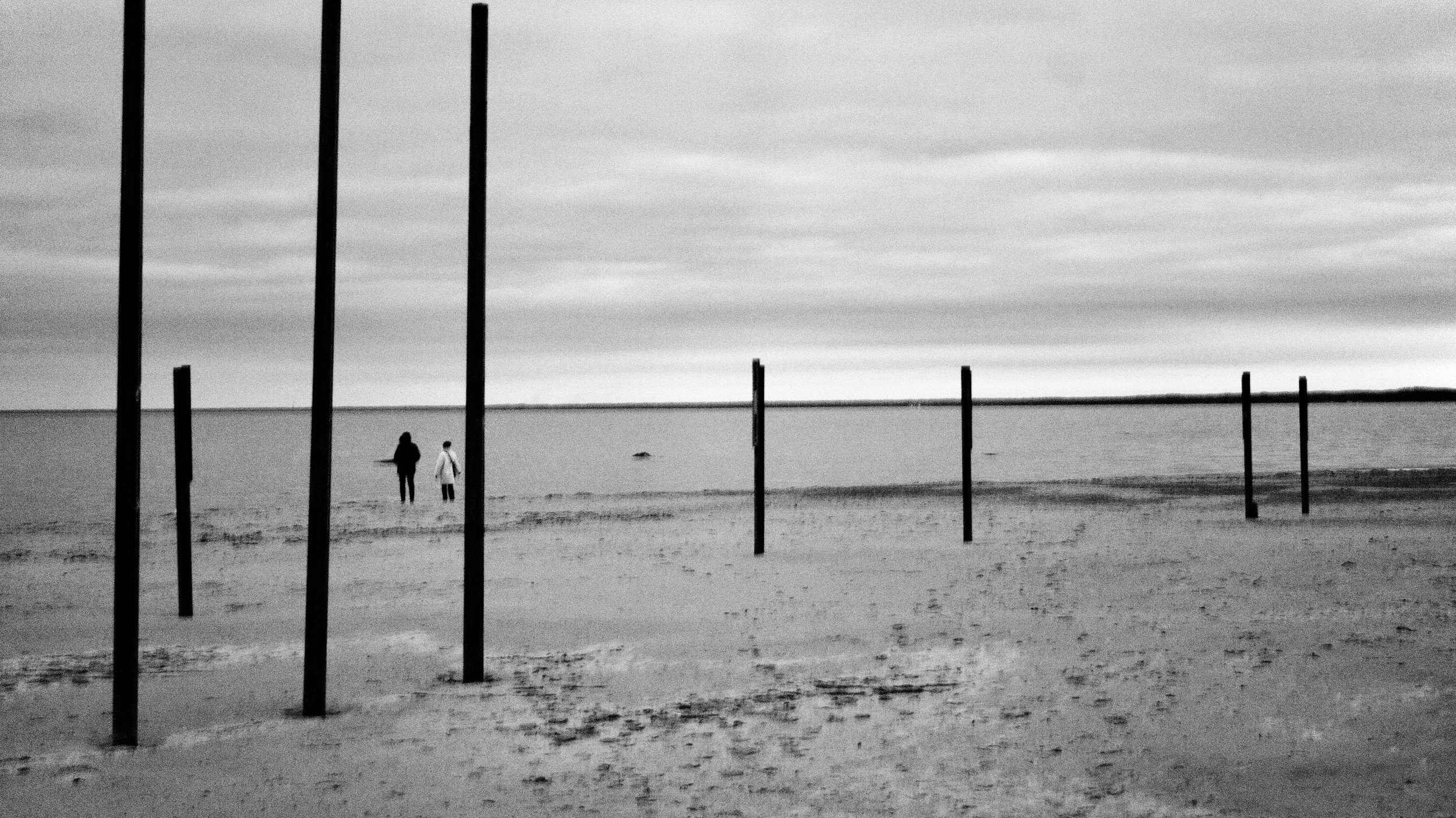 Black and white photo of two people walking on a beach near the water, with several wooden posts in the sand and overcast sky.