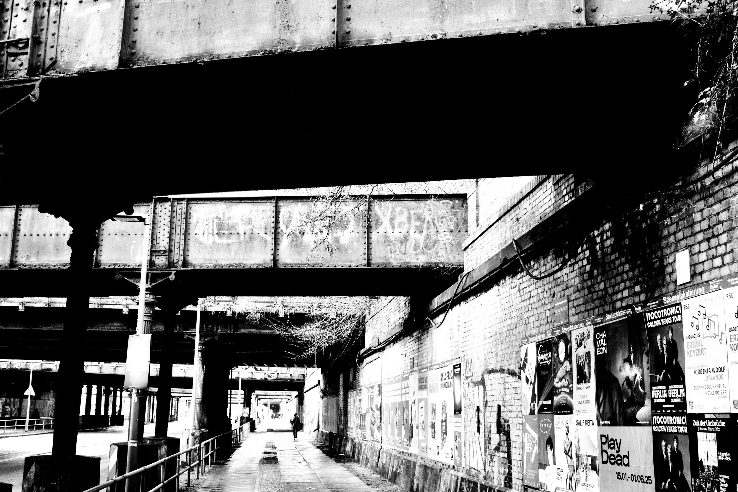 Black-and-white photo of an urban street scene under a bridge, with posters on a brick wall on the right side and a person walking under the bridge.
