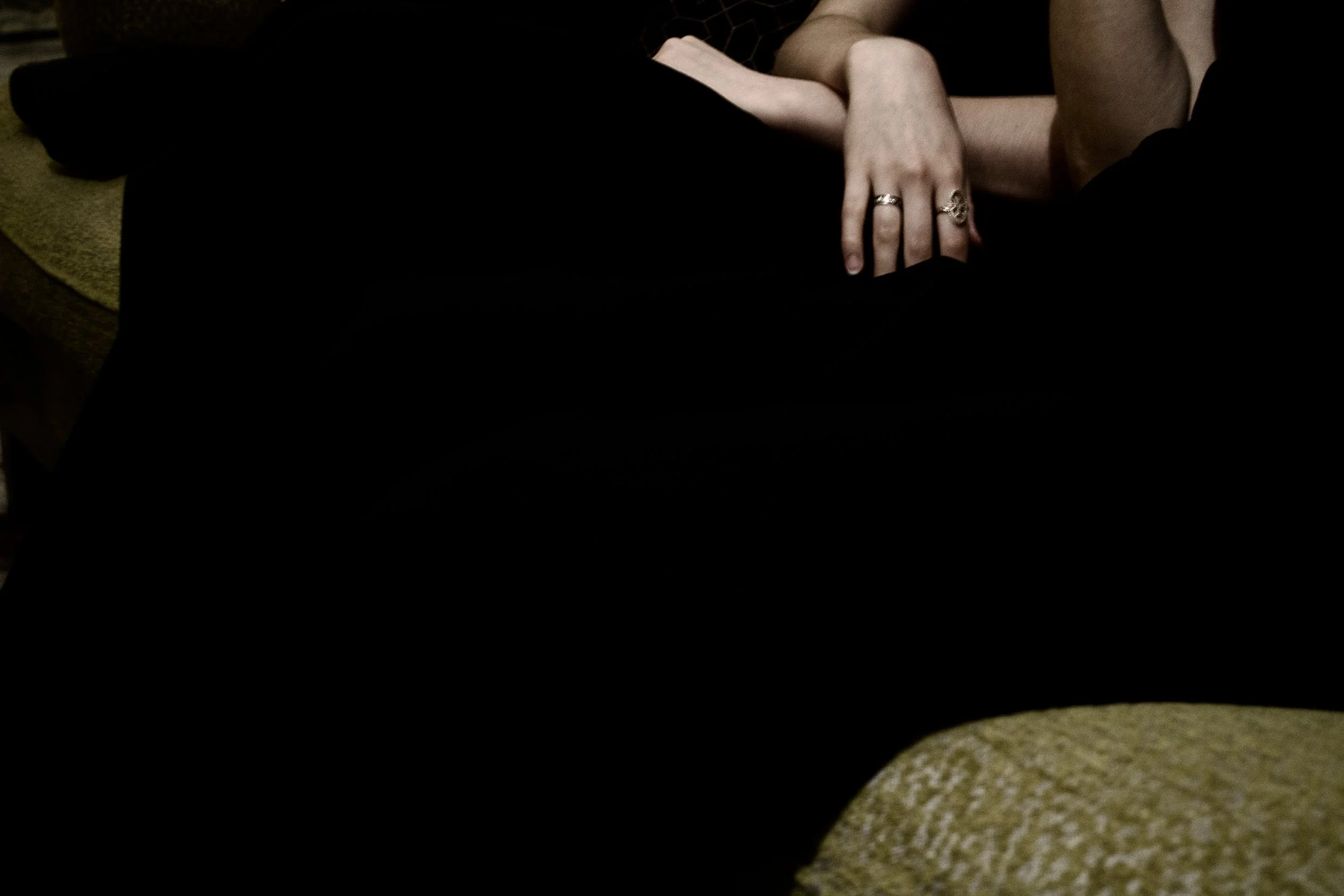 Close-up of a person's arm resting on their lap, showing fingers with rings, with part of their black clothing and beige shoes visible.