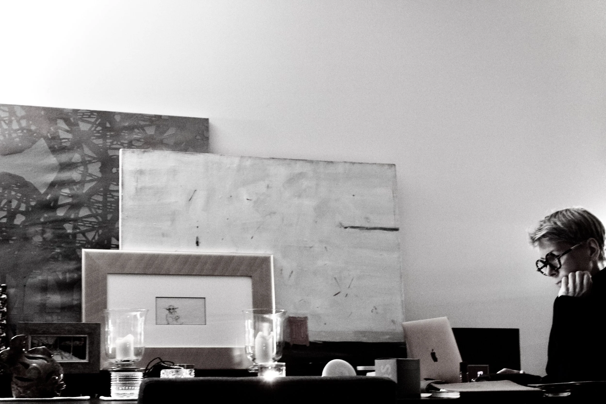 A black and white photo of a room with a woman wearing glasses on the right side, surrounded by framed pictures and decorative items on a table, with candles and glassware, and abstract artwork on the wall behind her.