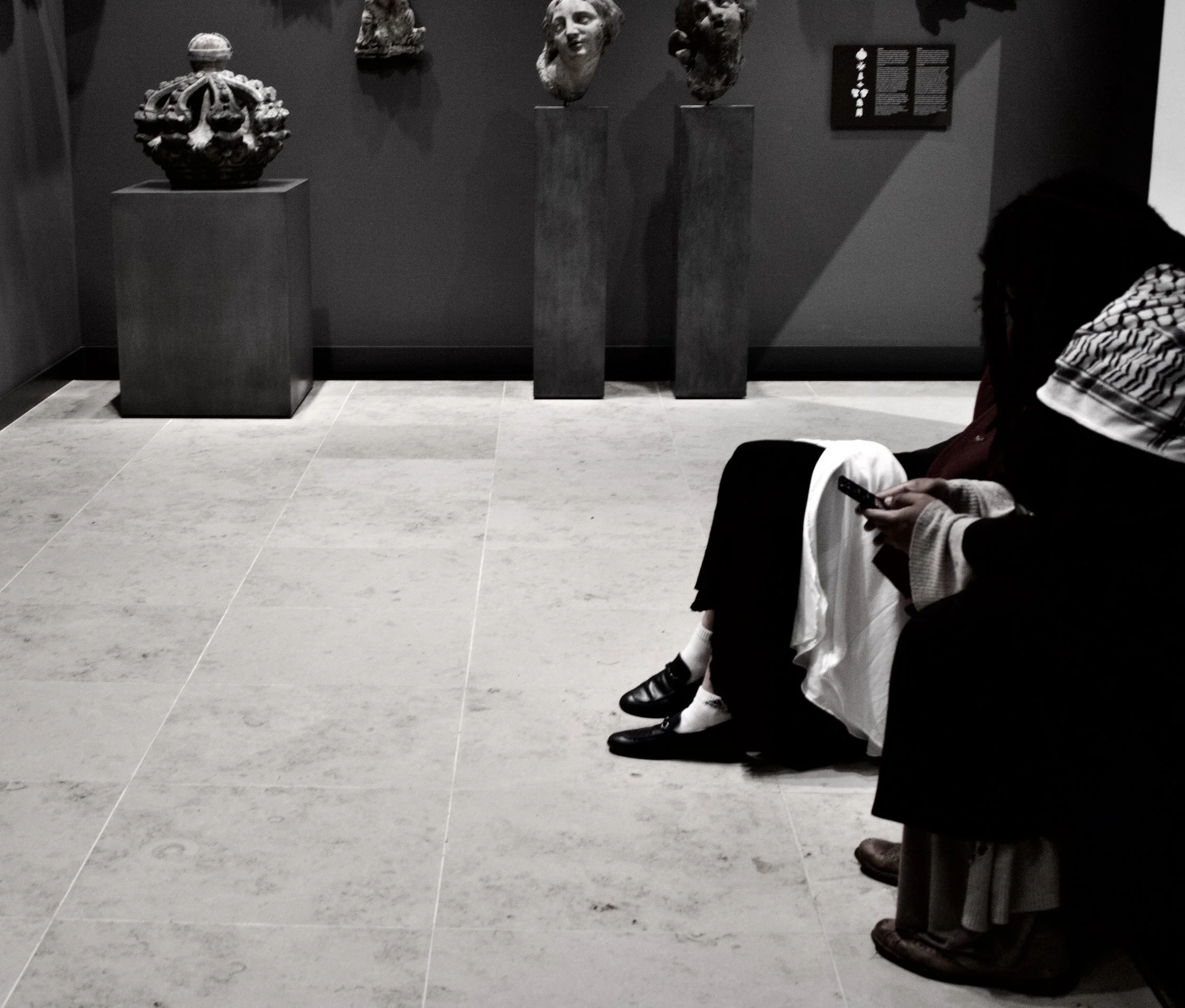 Museum exhibit with three sculptures of human heads on black pedestals and one ornate sculpture of a crown or headdress on a stand. A person in the foreground is sitting and looking at their phone; another person is partially visible on the right.