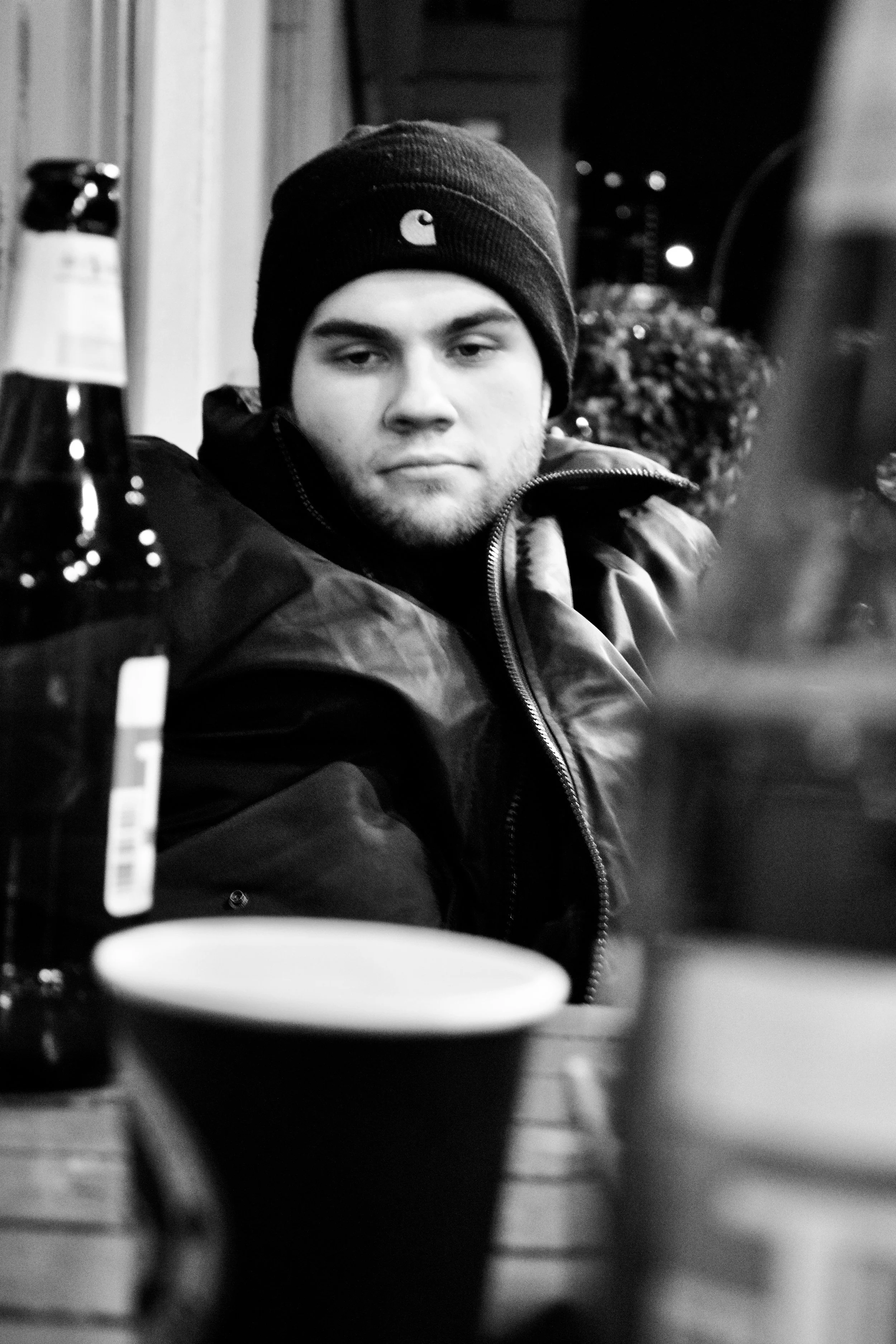 Black and white photo of a young man wearing a beanie hat and jacket, sitting in what appears to be a dimly lit indoor setting. The photo is taken from a table with a bottle, a mug, and other objects in the foreground.