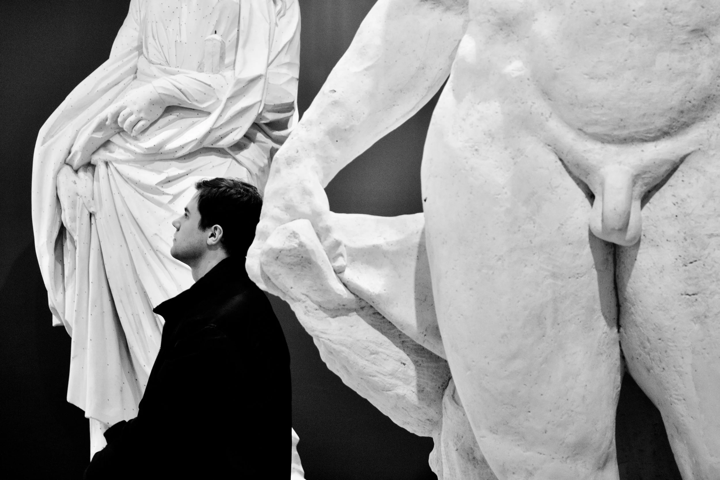 A man with dark hair looking at a large classical marble sculpture of a woman with draped clothing. The sculpture's arm is extended downward and the texture of the marble is visible.