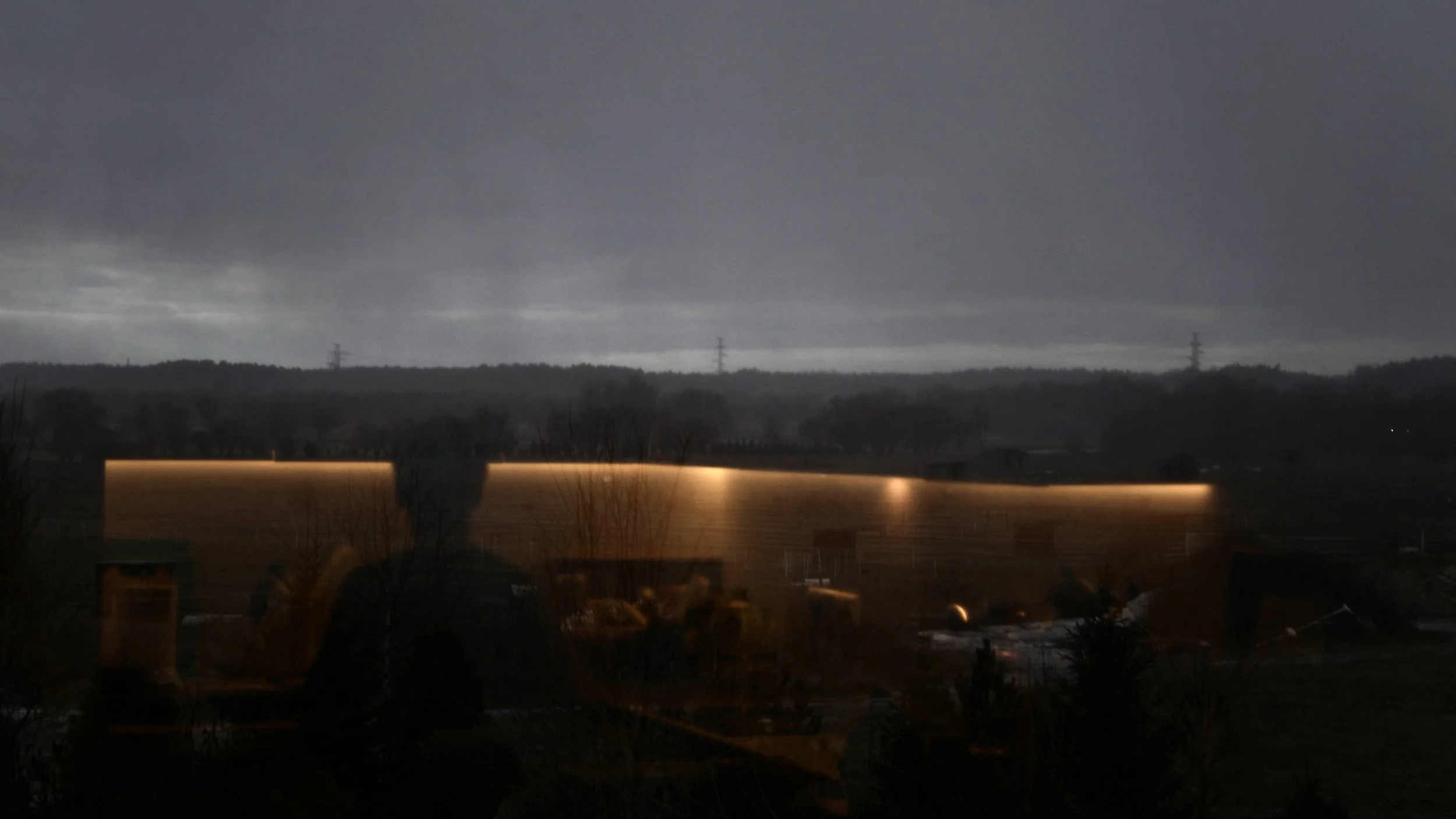 Dark cloudy sky over a distant landscape with hills, power lines, and illuminated building edges in the foreground.