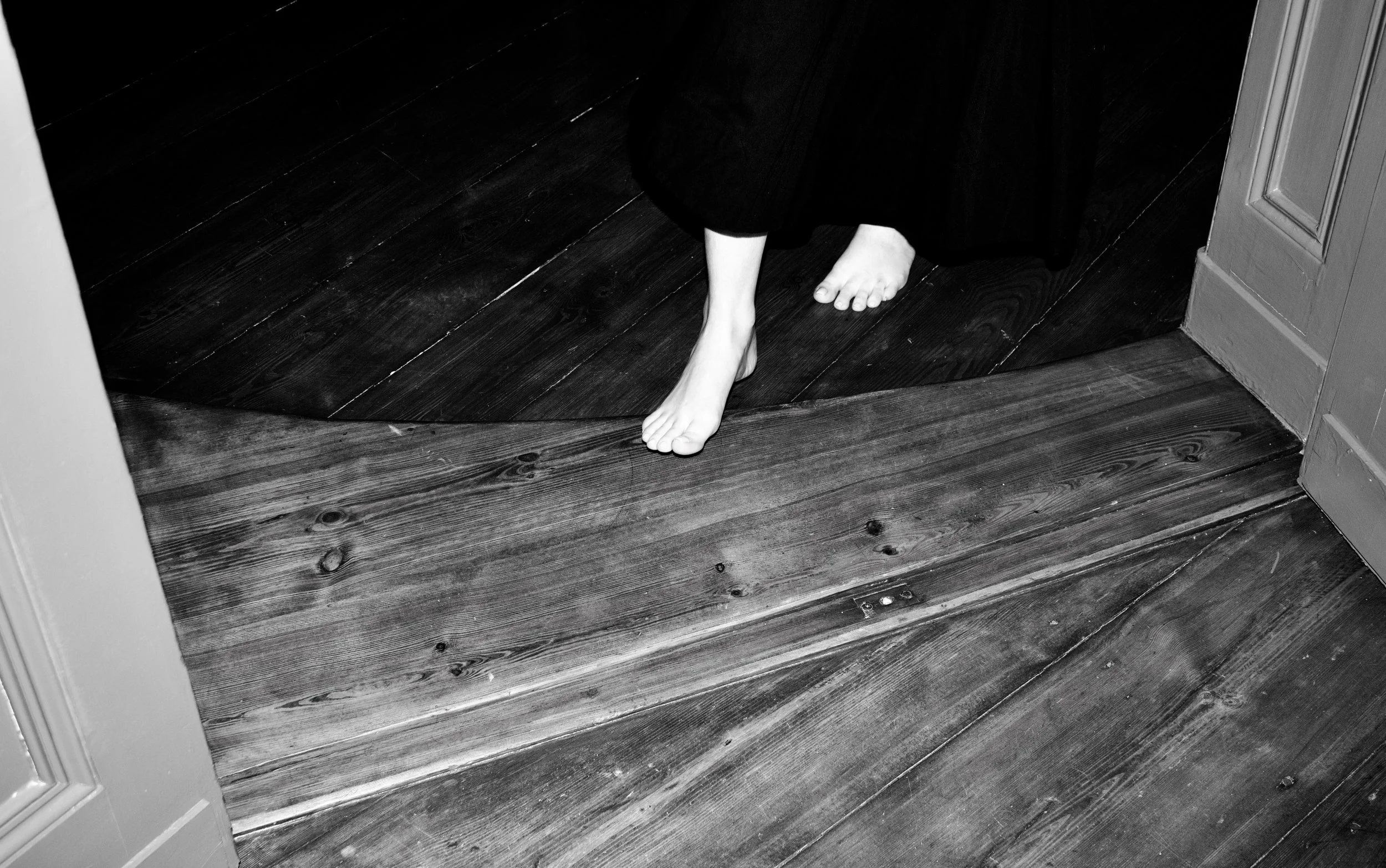 A person walking barefoot on wooden floors, wearing a long skirt or dress, with part of a door frame visible on the right side of the image.