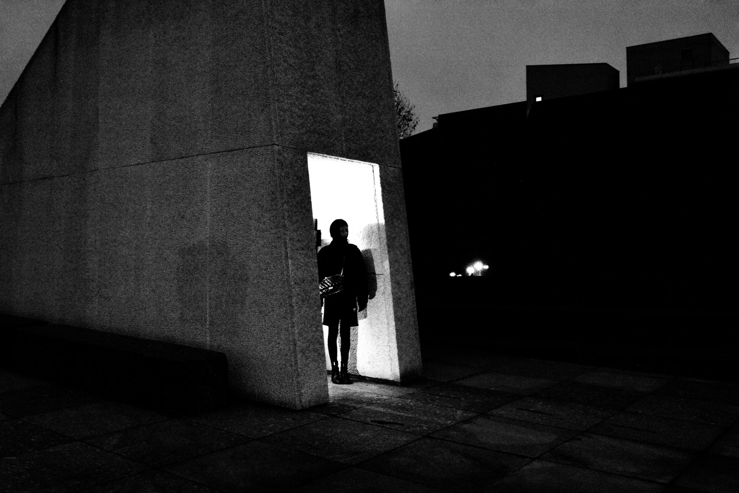 A person standing in a doorway at night, illuminated from behind, with a dark urban background and modern buildings silhouetted against the night sky.