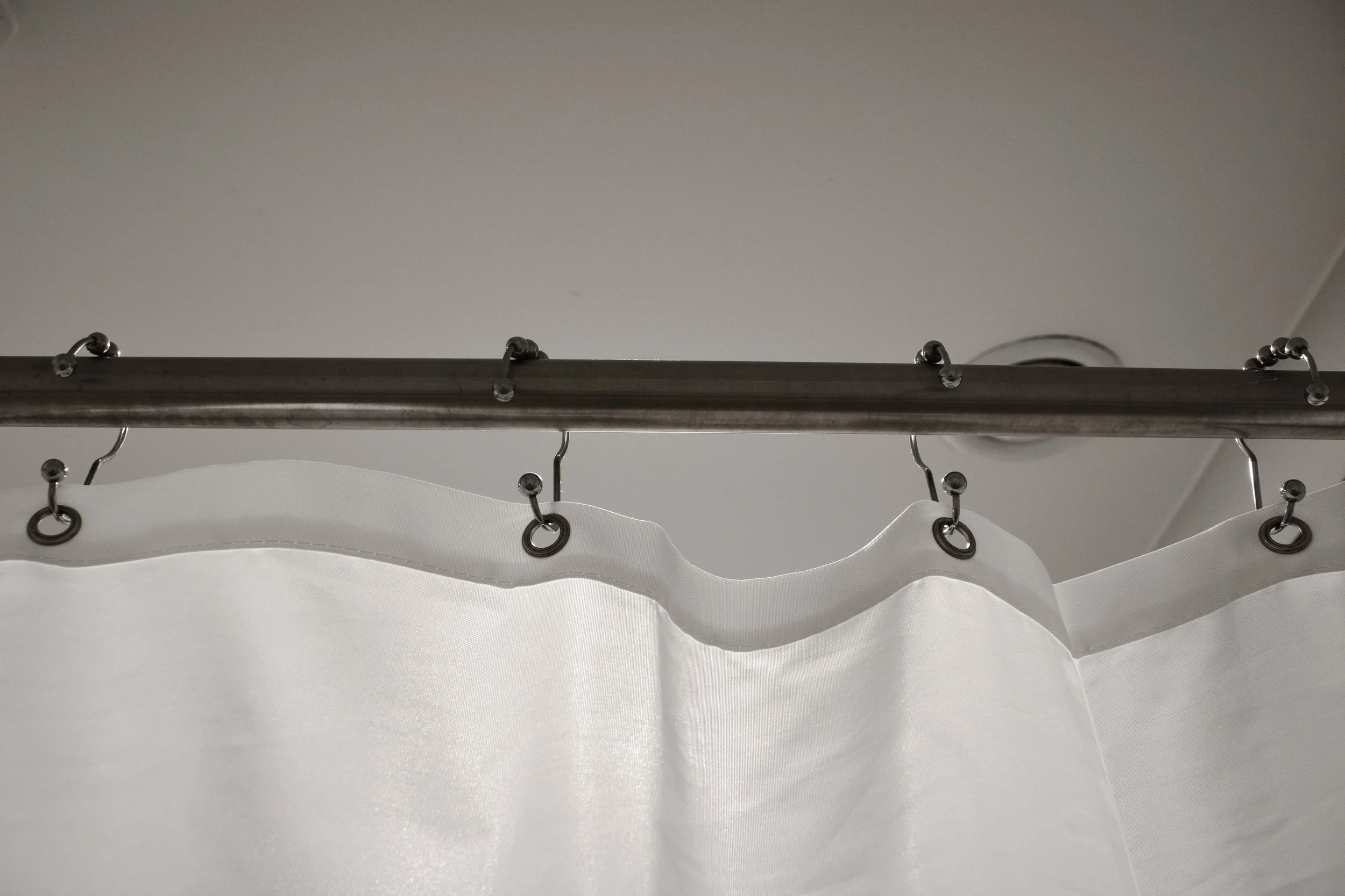 A metal curtain rod with white fabric curtains hanging from it, attached with metal hooks.