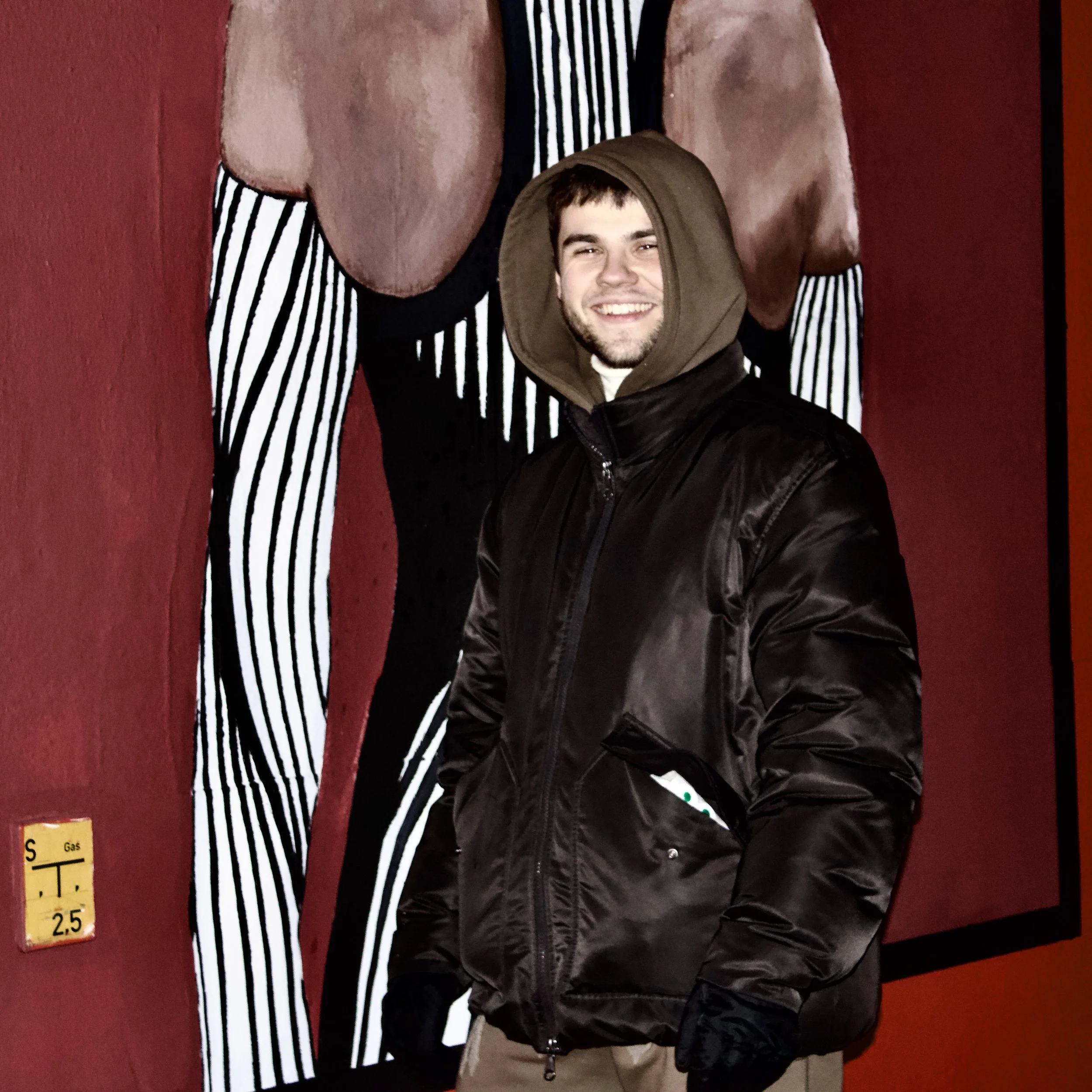 A young man with short hair and a beard smiling while wearing a brown hooded jacket, standing against a red wall with a large abstract art piece behind him.