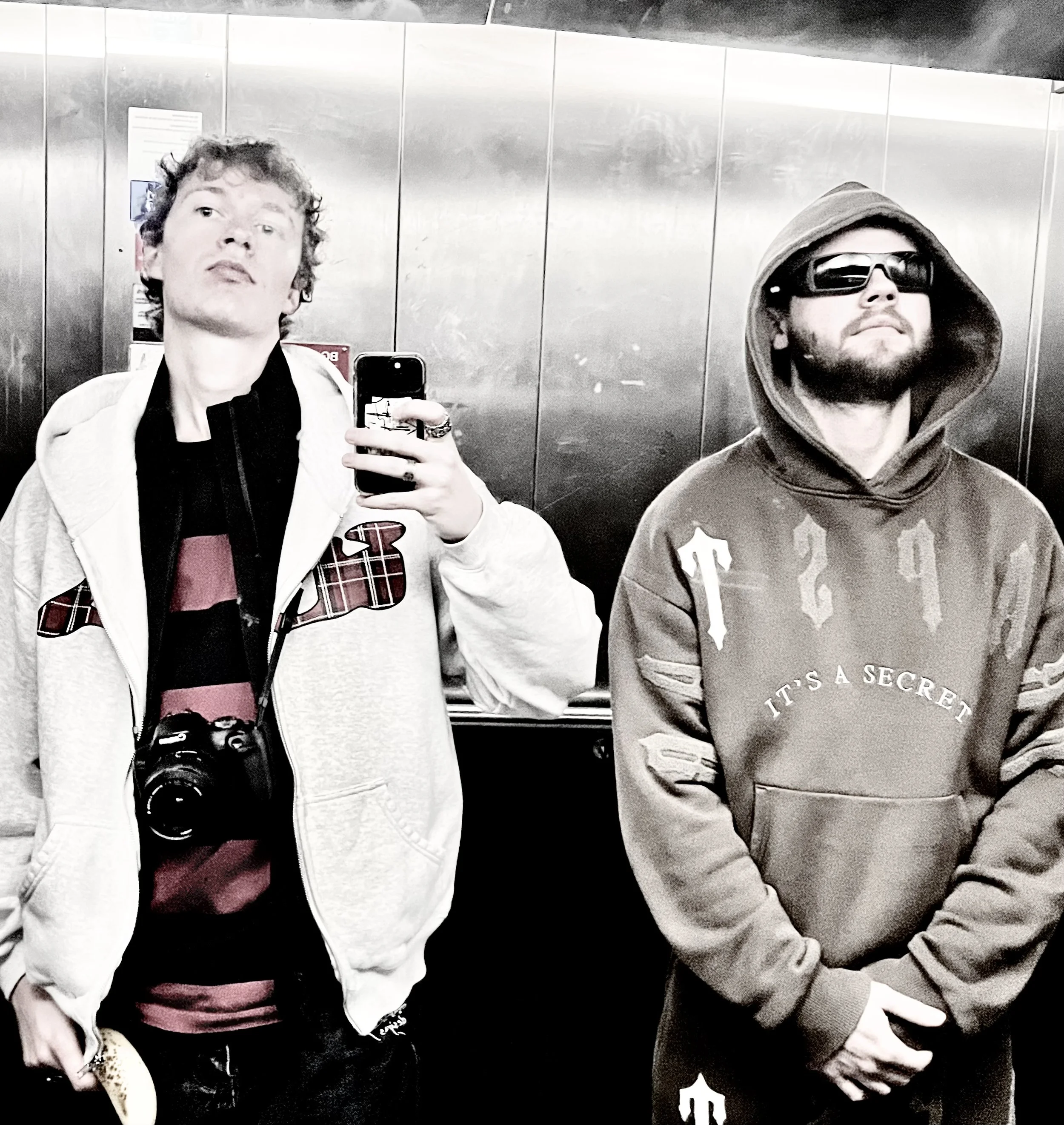 Two men taking a selfie in a reflective elevator. The man on the left has curly hair, is wearing a white hoodie and a red and black shirt underneath, and has a camera hanging around his neck. The man on the right has a beard, is wearing a hoodie with the hood up, and sunglasses. The background shows metal panels of the elevator interior.