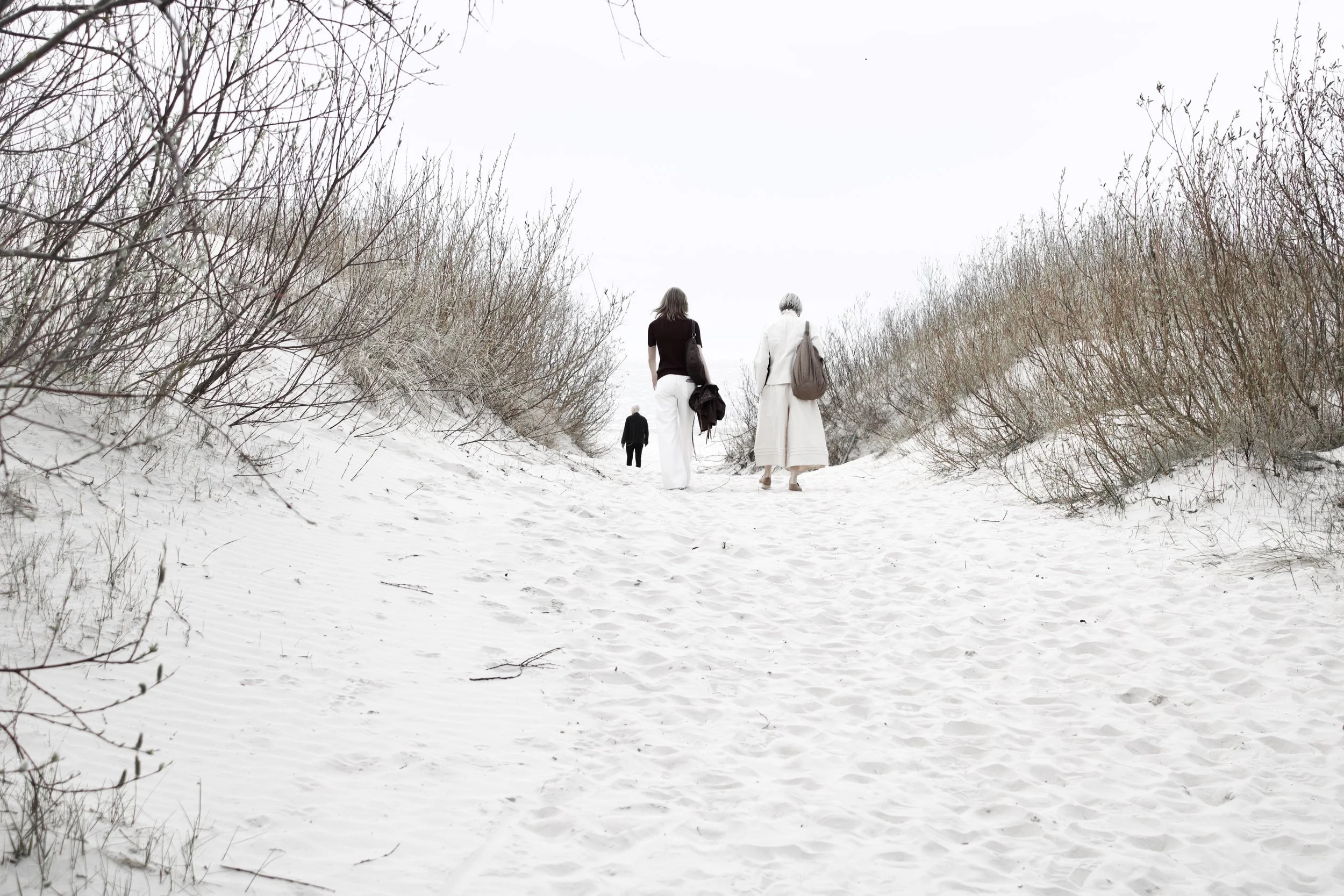 Two women walking through a snow-covered path with bare bushes on either side, carrying handbags and dressed in winter clothing.