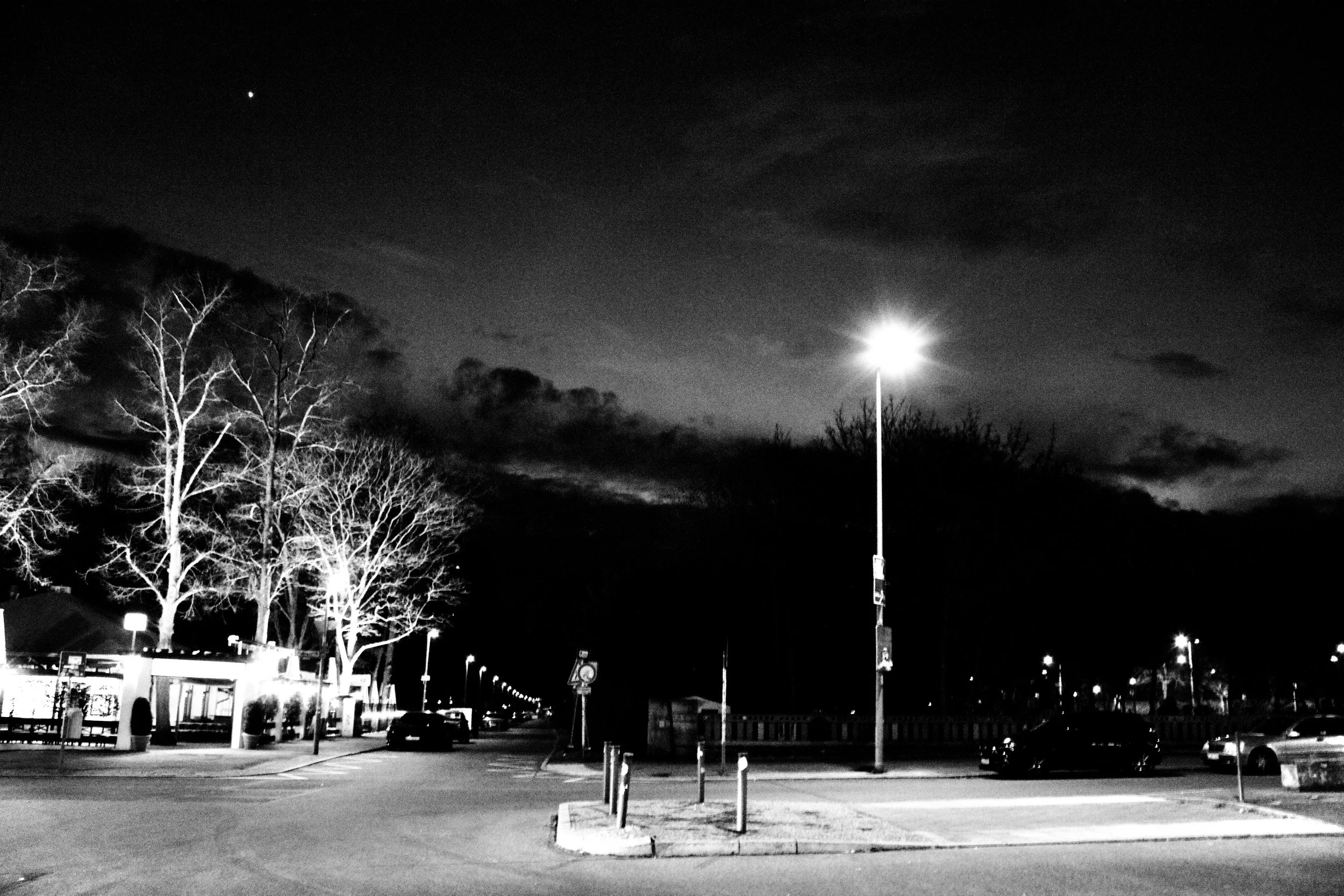 Nighttime scene of a parking lot with a few cars, leafless trees, a bright streetlamp, and a dark sky with some clouds and a visible star.