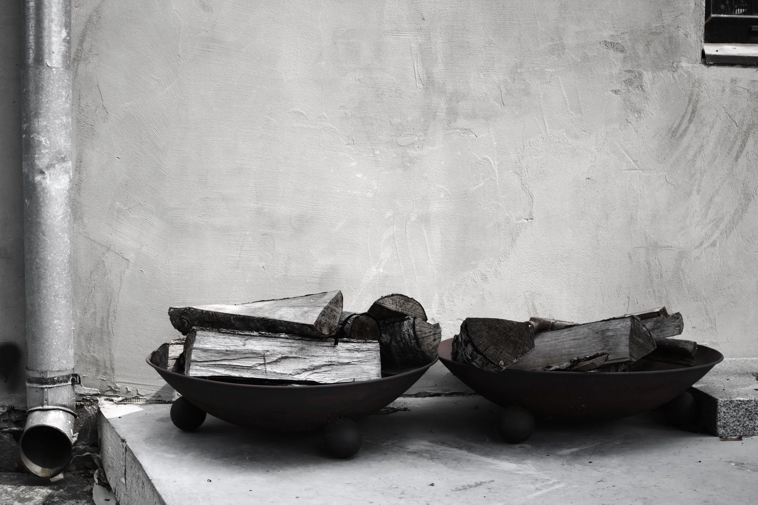 Firewood logs stacked in black bowls in front of a white plastered wall with a grey drainpipe on the left.