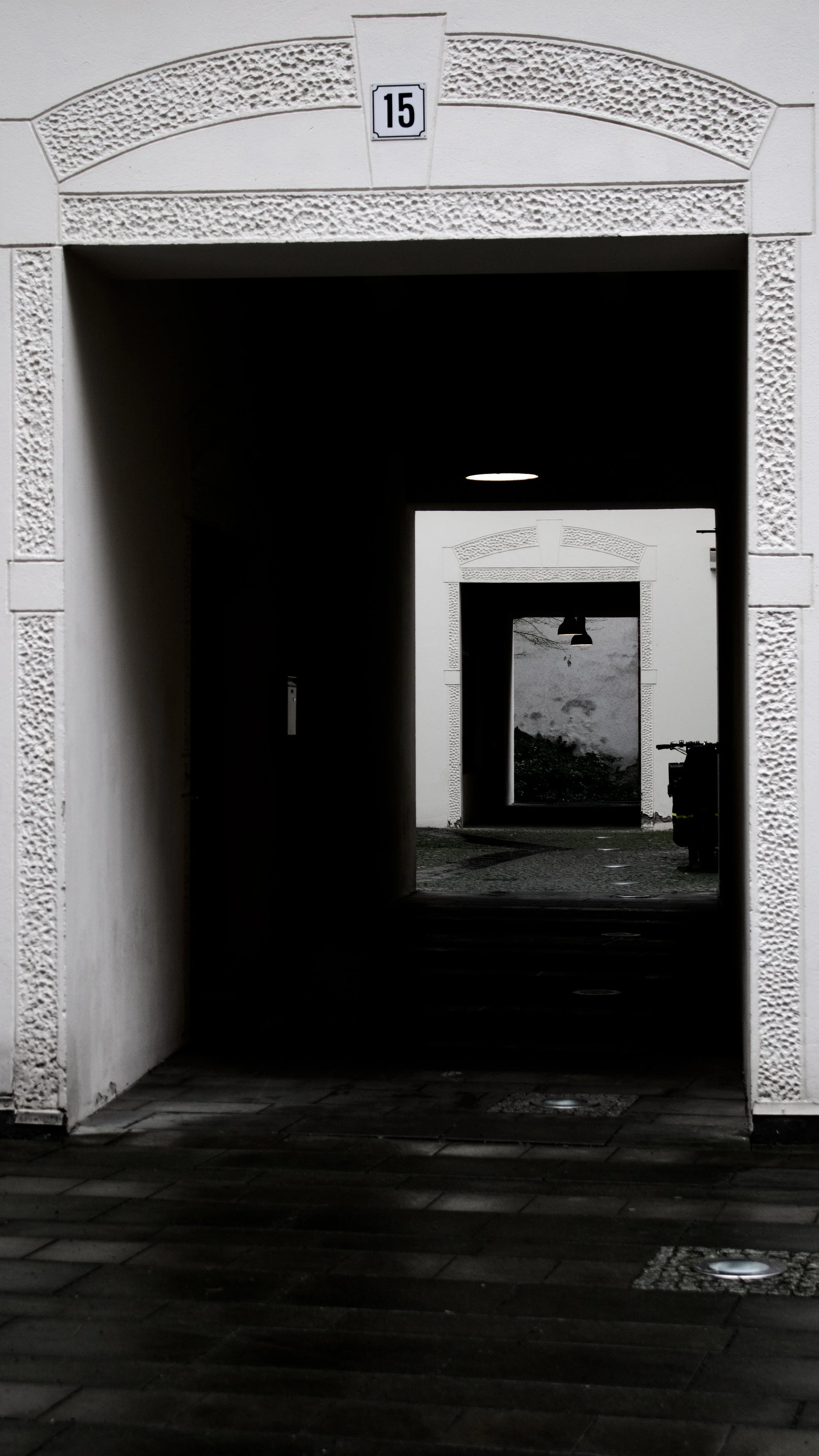 A series of white archways with textured borders creating a tunnel effect, with the closest archway displaying the number 15 on a sign, leading to an outdoor area with dark ground and a distant white wall.