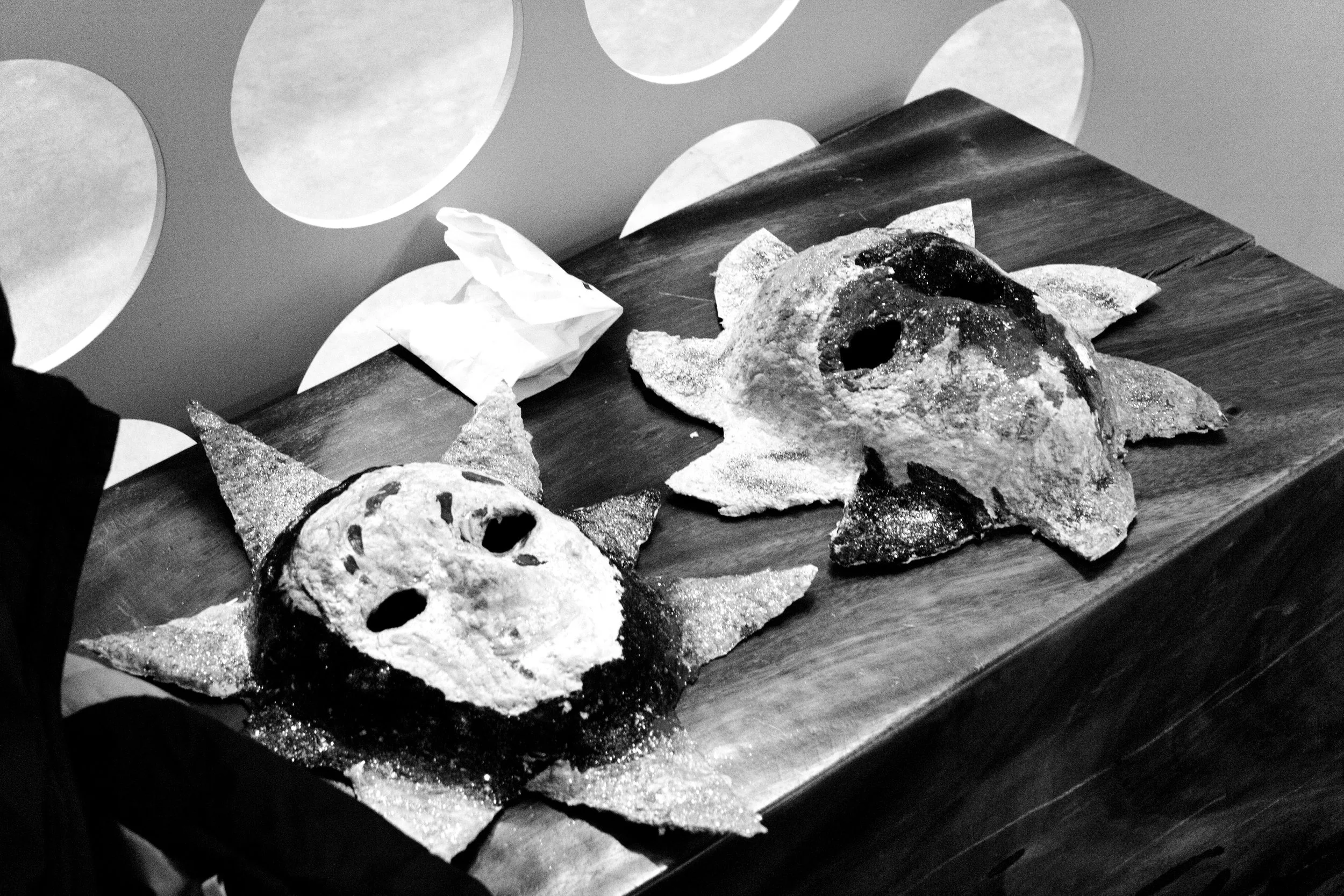 Black and white photo of several masks made from bread and other items, arranged on a wooden surface with a light-colored wall in the background.