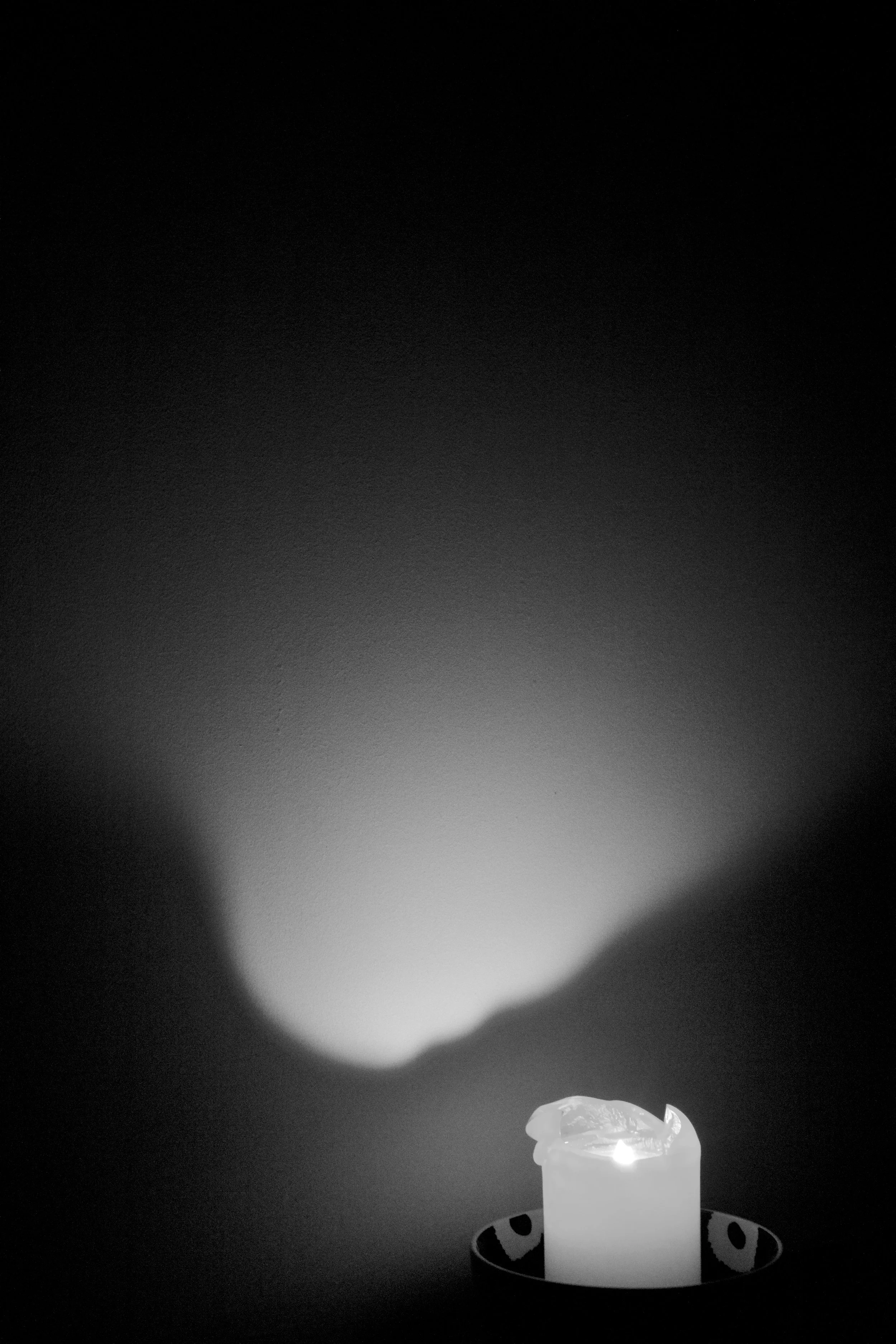 A white candle in a decorative holder, burning with a bright flame, casts a glow and shadow on a dark background.
