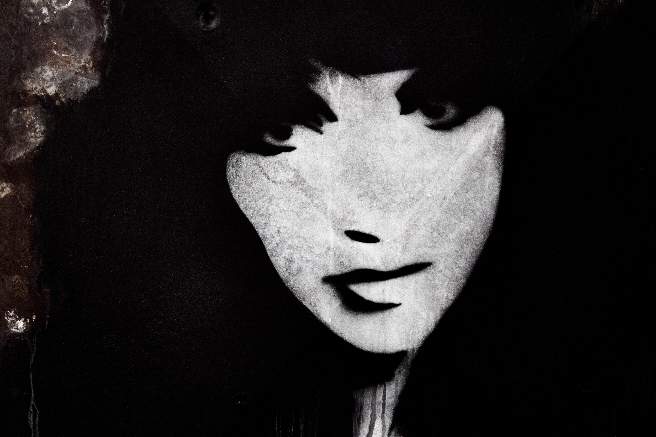 A black and white photo of a person wearing a white mask, with dark hair and eyes visible above the mask, against a dark background.