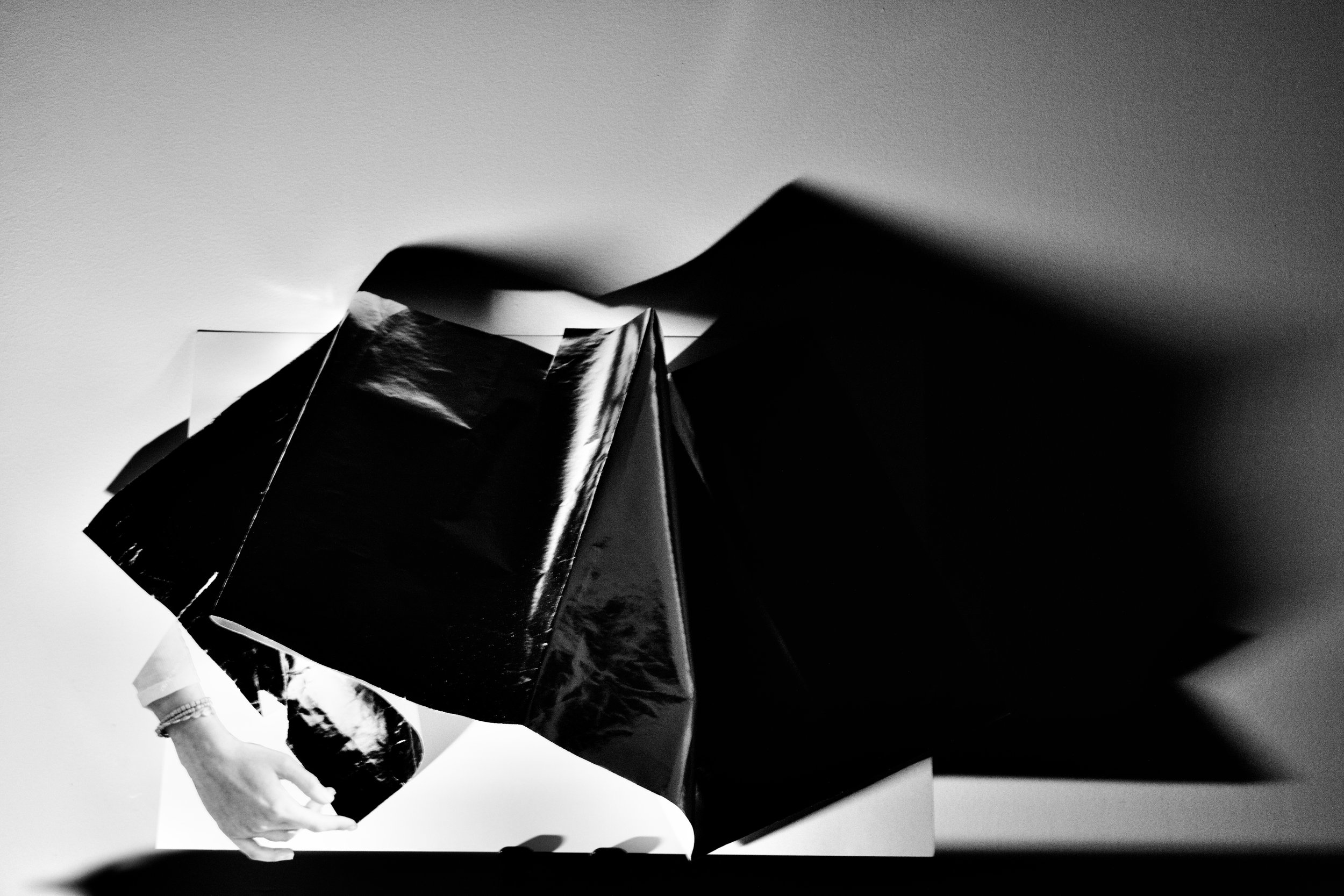 A person with a bracelet on their wrist holding a piece of paper or photograph, with an abstract inverted image above, involving geometric shapes and shadows.