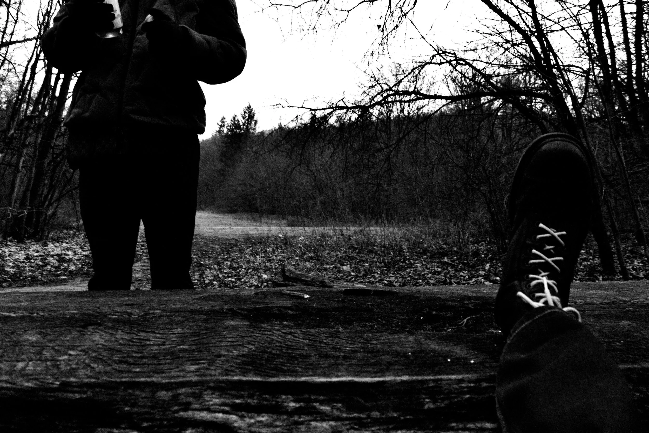 Black and white photograph of a person standing on a path in a wooded area during the day, with trees and branches overhead. The person is wearing dark clothing and holding a beverage can. A person's foot wearing a sneaker is visible in the foregroun