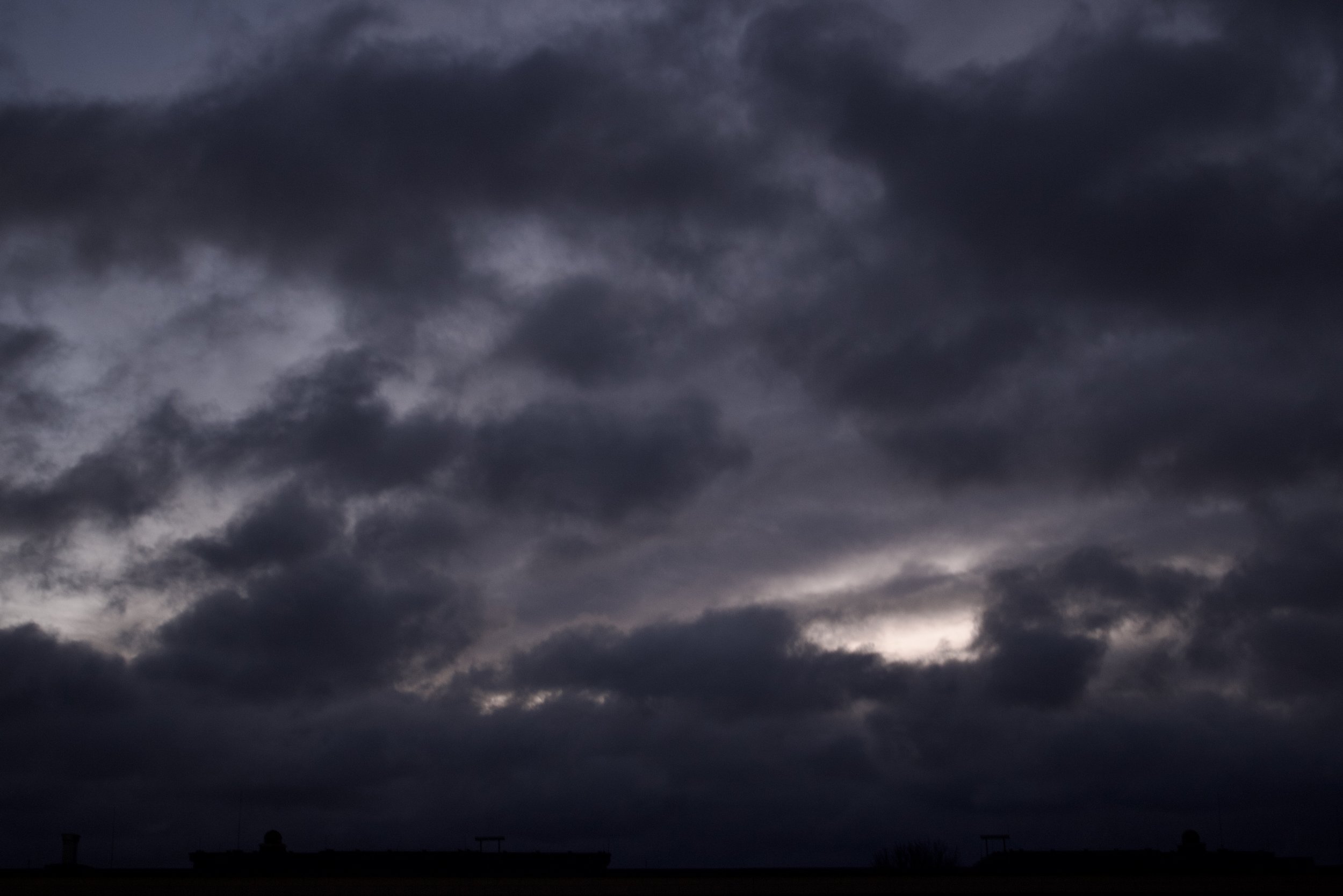 Dark, cloudy storm sky with a faint hint of light near the horizon, and silhouettes of structures at the bottom.