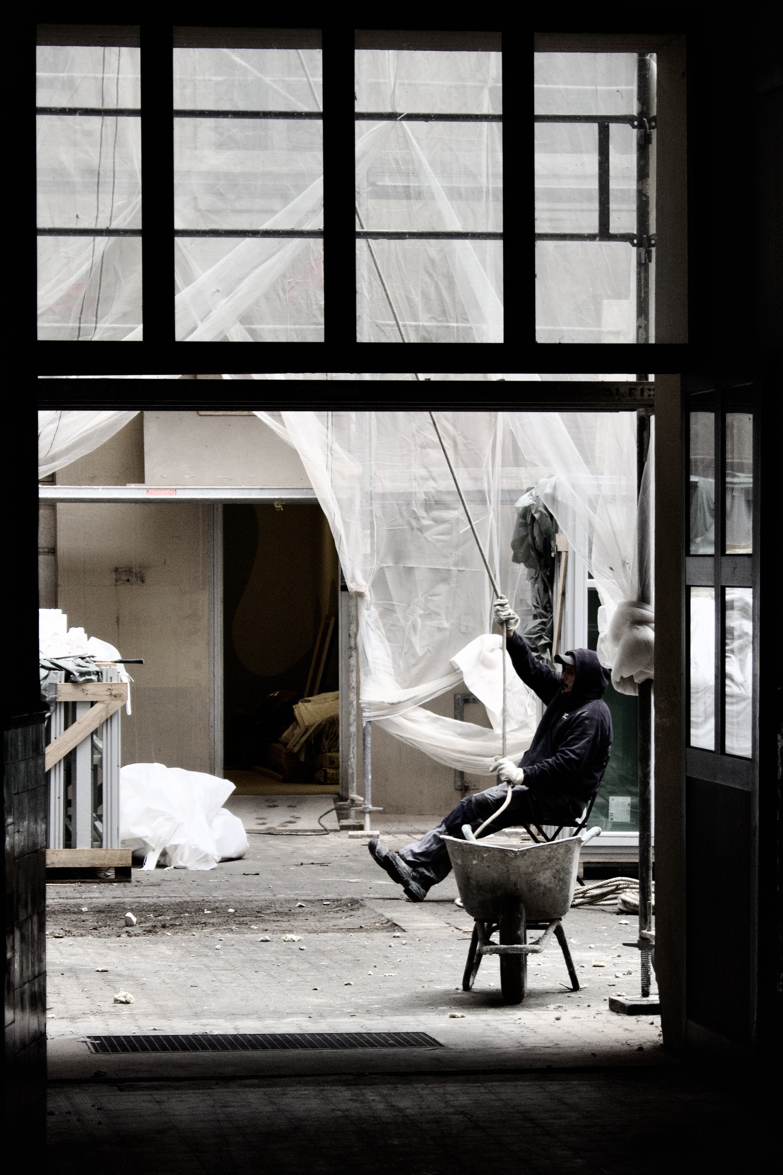 A worker sitting on a chair inside a building, holding a rope, with construction materials and plastic sheeting visible through large window openings.