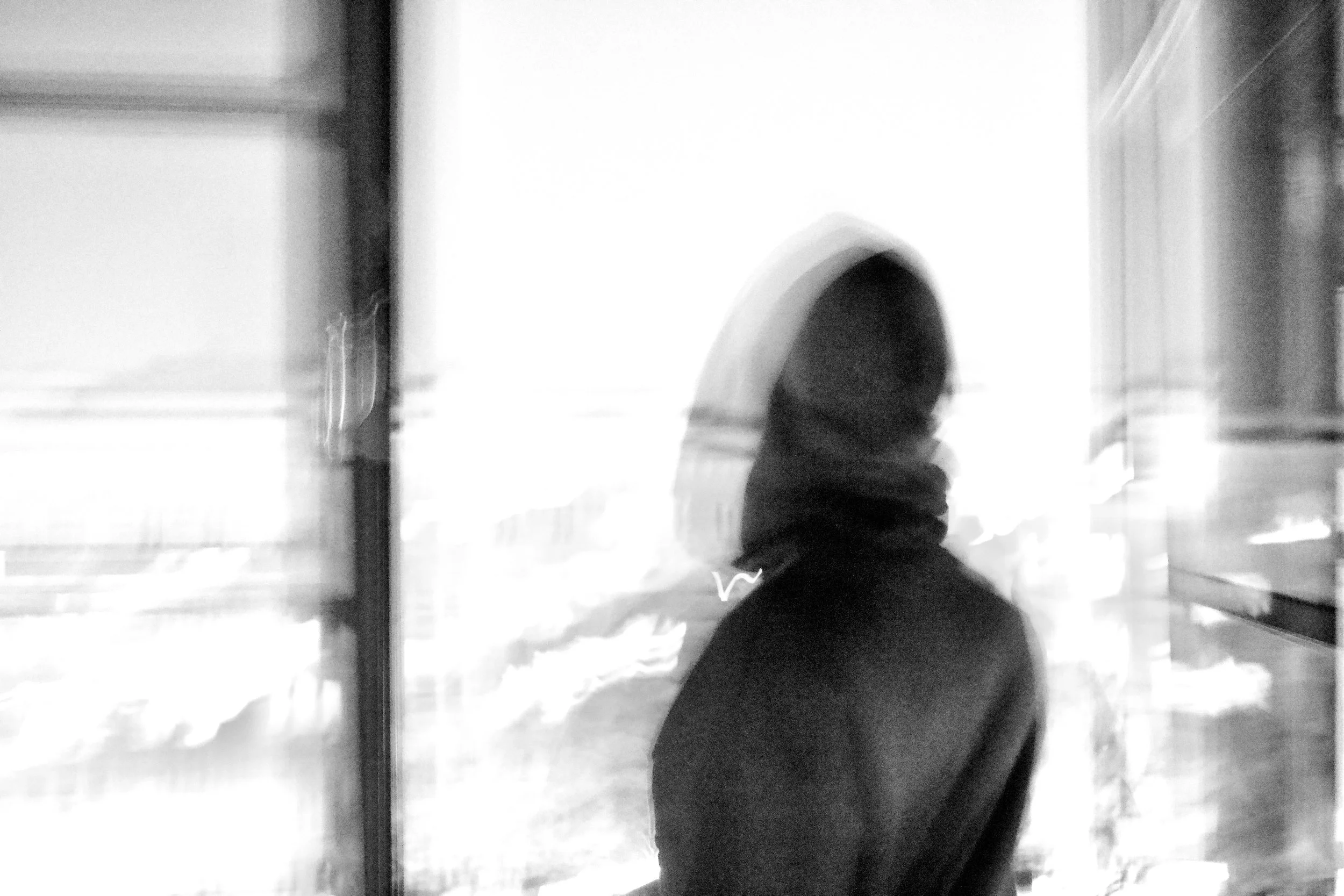 A black and white photograph of a person with curly hair walking past a glass window or door, with blurred reflections of the outside visible on the surface.