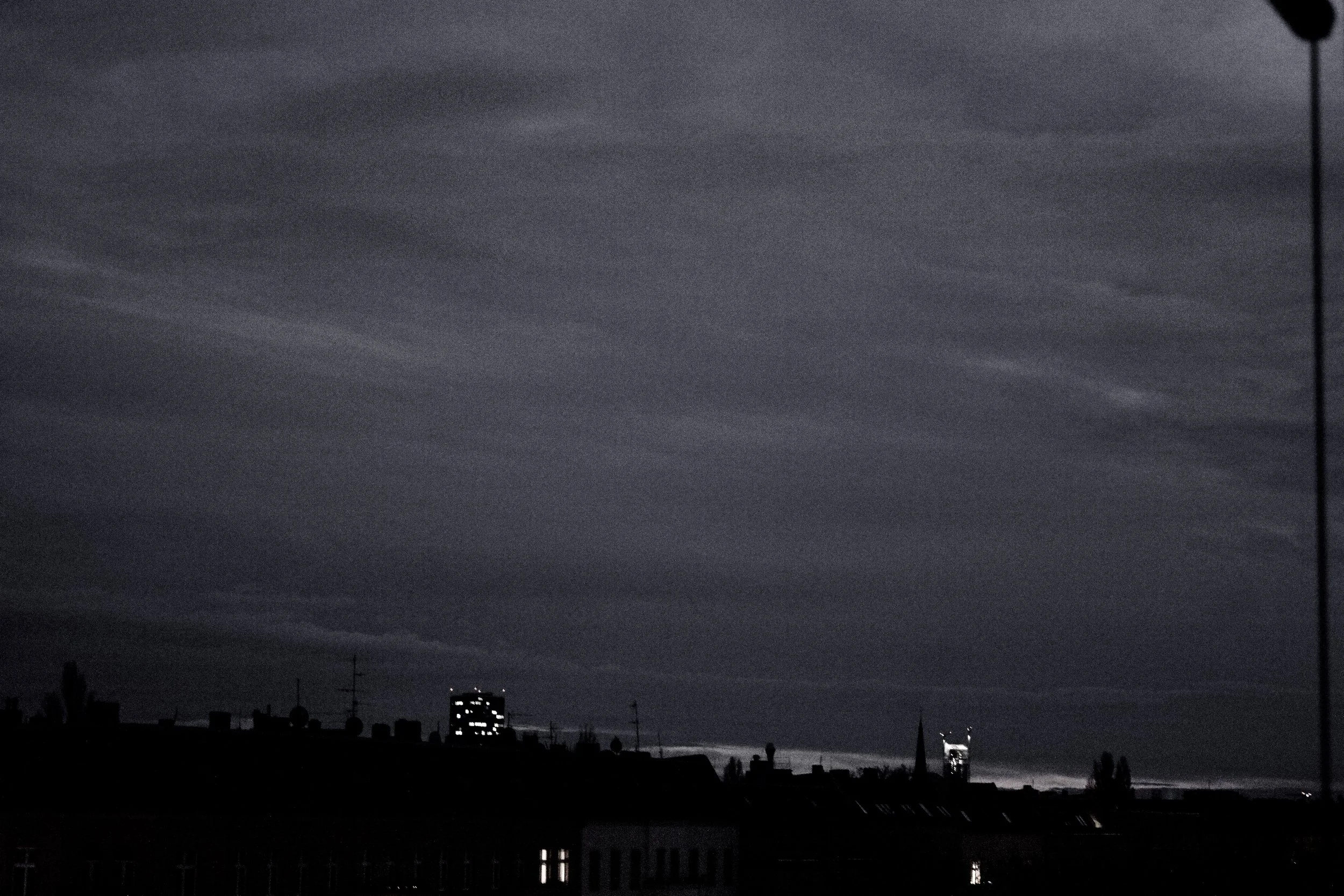 Dark, cloudy sky over a cityscape with silhouetted buildings and rooftops, some with antennas. A few city lights are visible amid the dark scene.