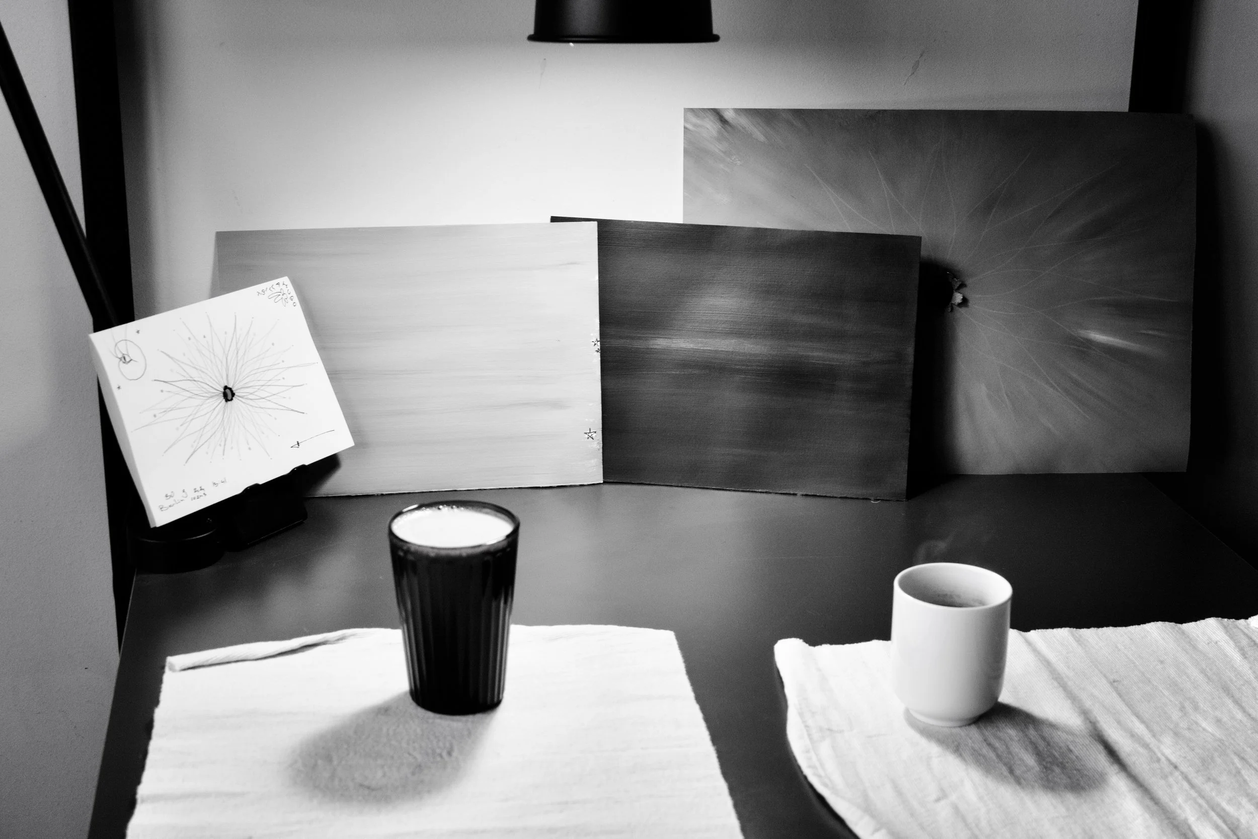 Black and white photo of a dining table with three cups, a napkin, and a glass. Behind the table are three boards or panels leaning against the wall with a note or drawing on one of them, illuminated by a hanging lamp.