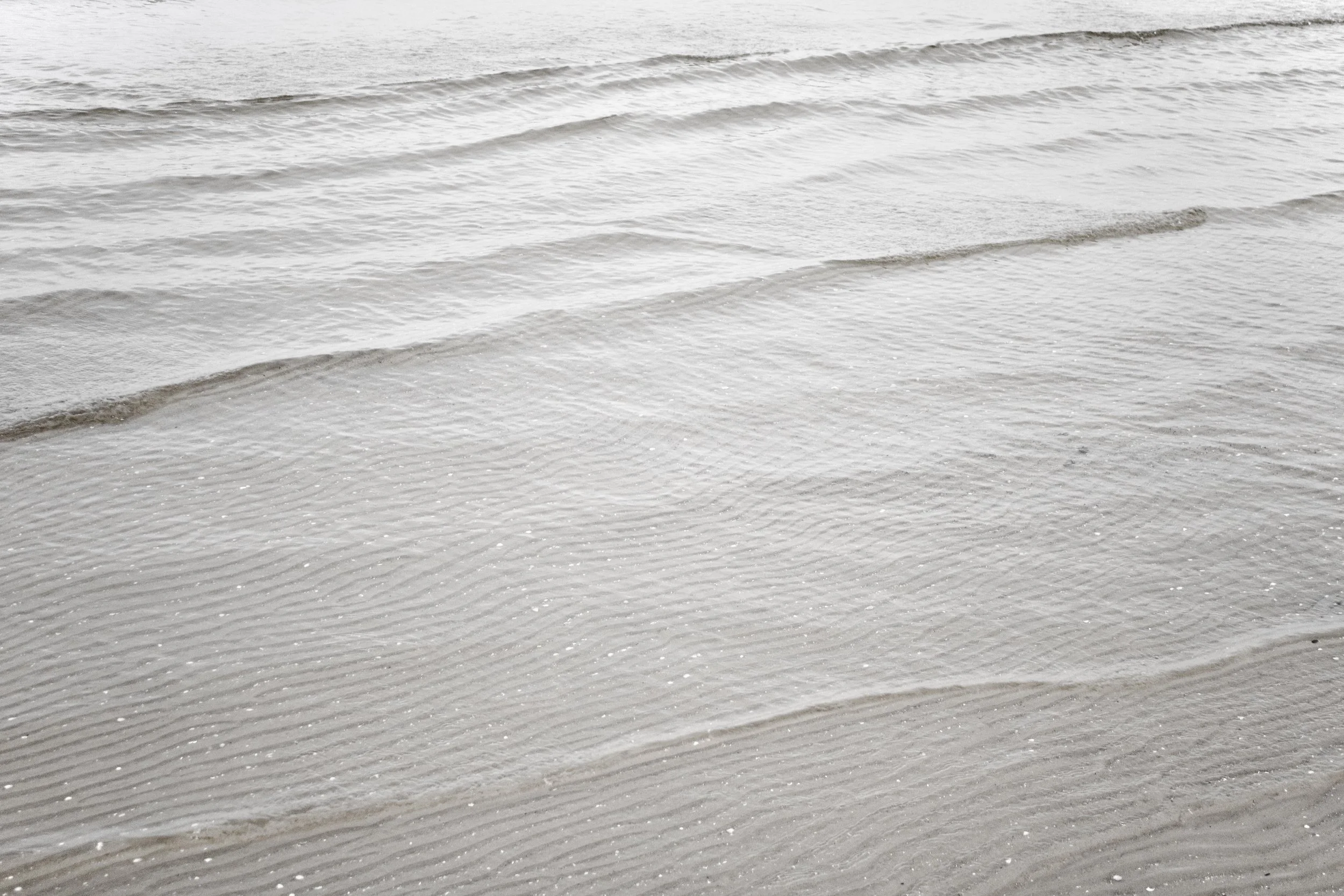 Close-up of wet sand on a beach with small, gentle waves in the water.