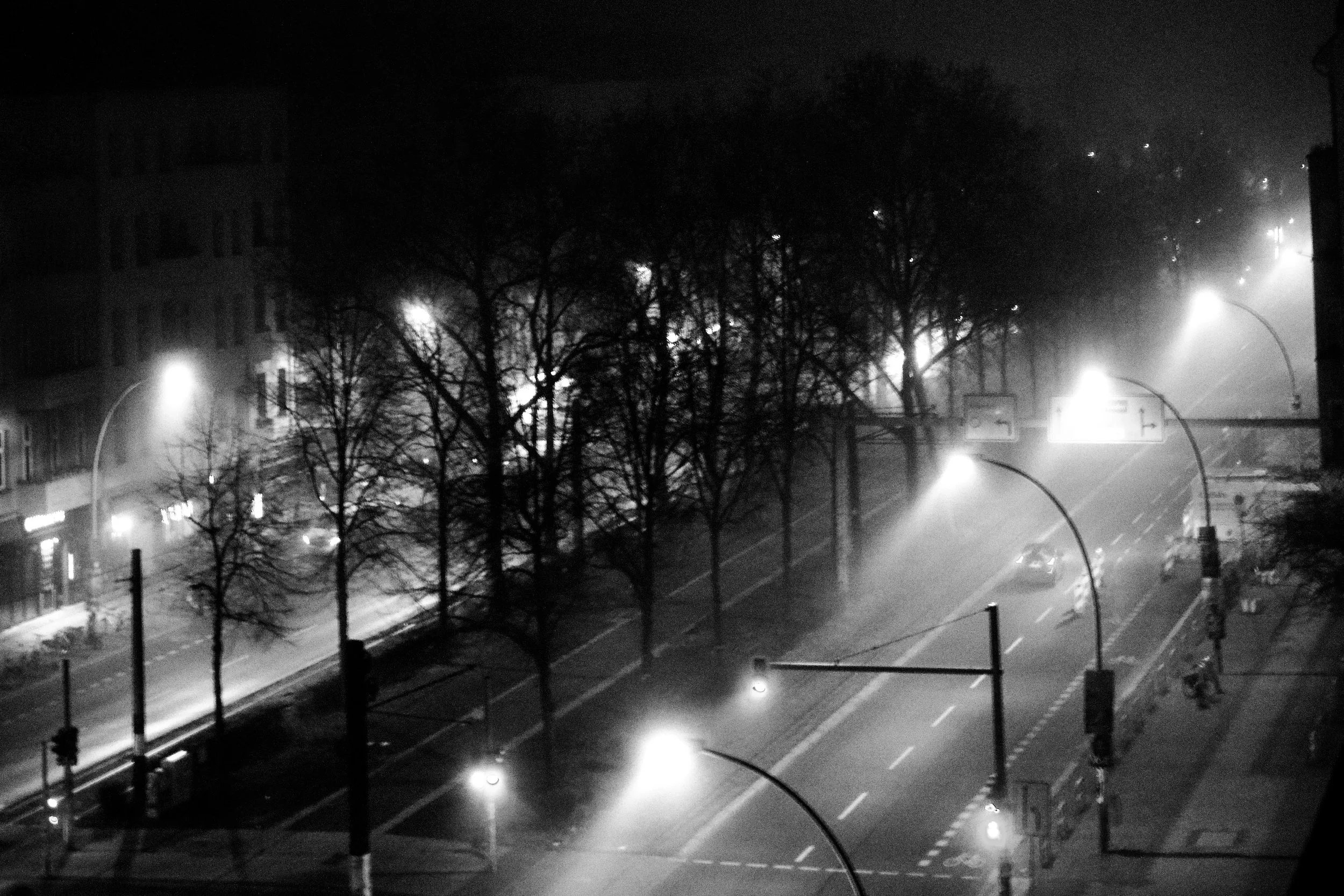 A black and white photo of a foggy city street at night with a few cars and illuminated streetlights. There are trees without leaves and apartment buildings in the background.