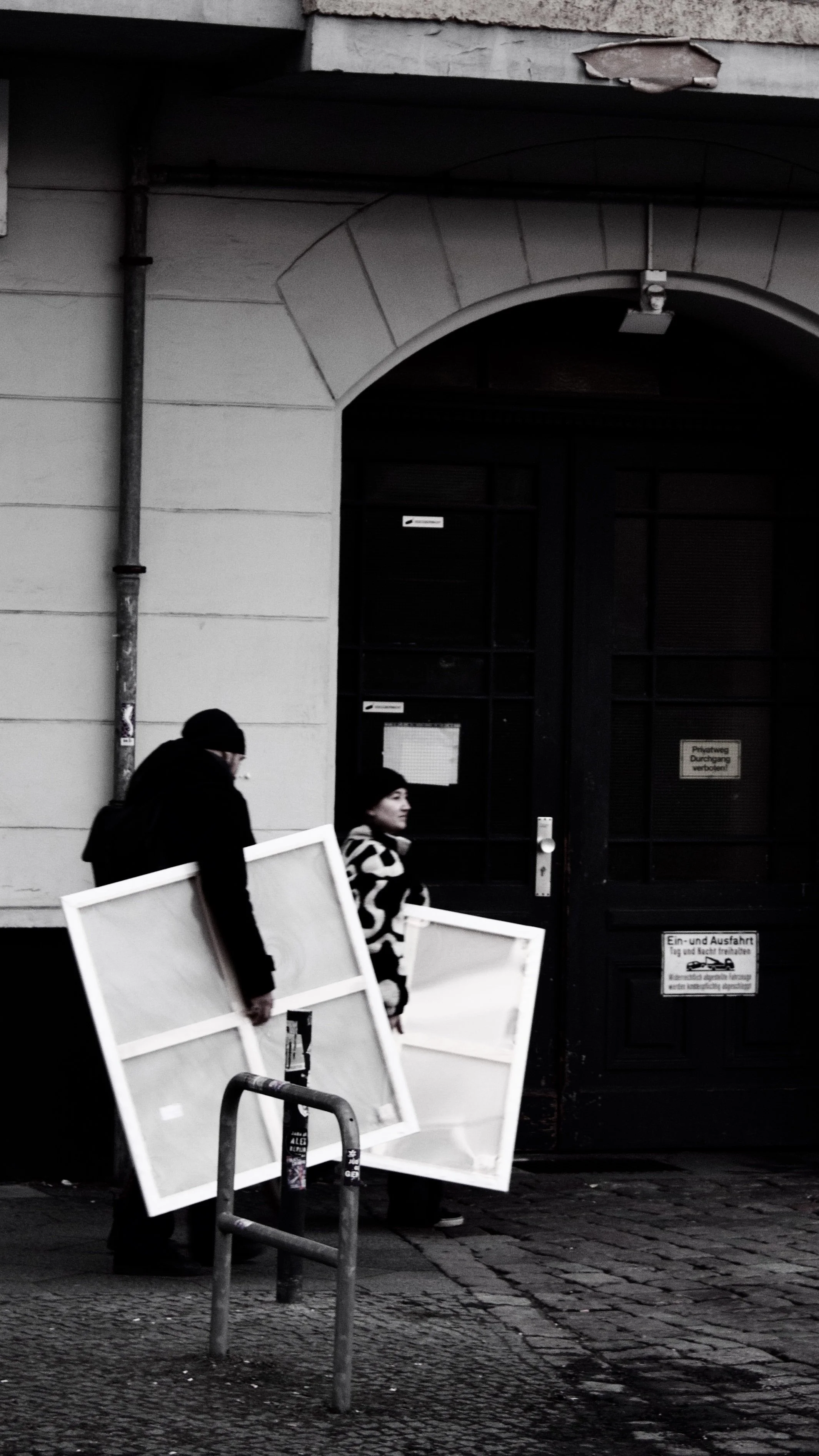 Two people walking past a door, carrying white frames, on a city street in front of a building with signs and a black door.