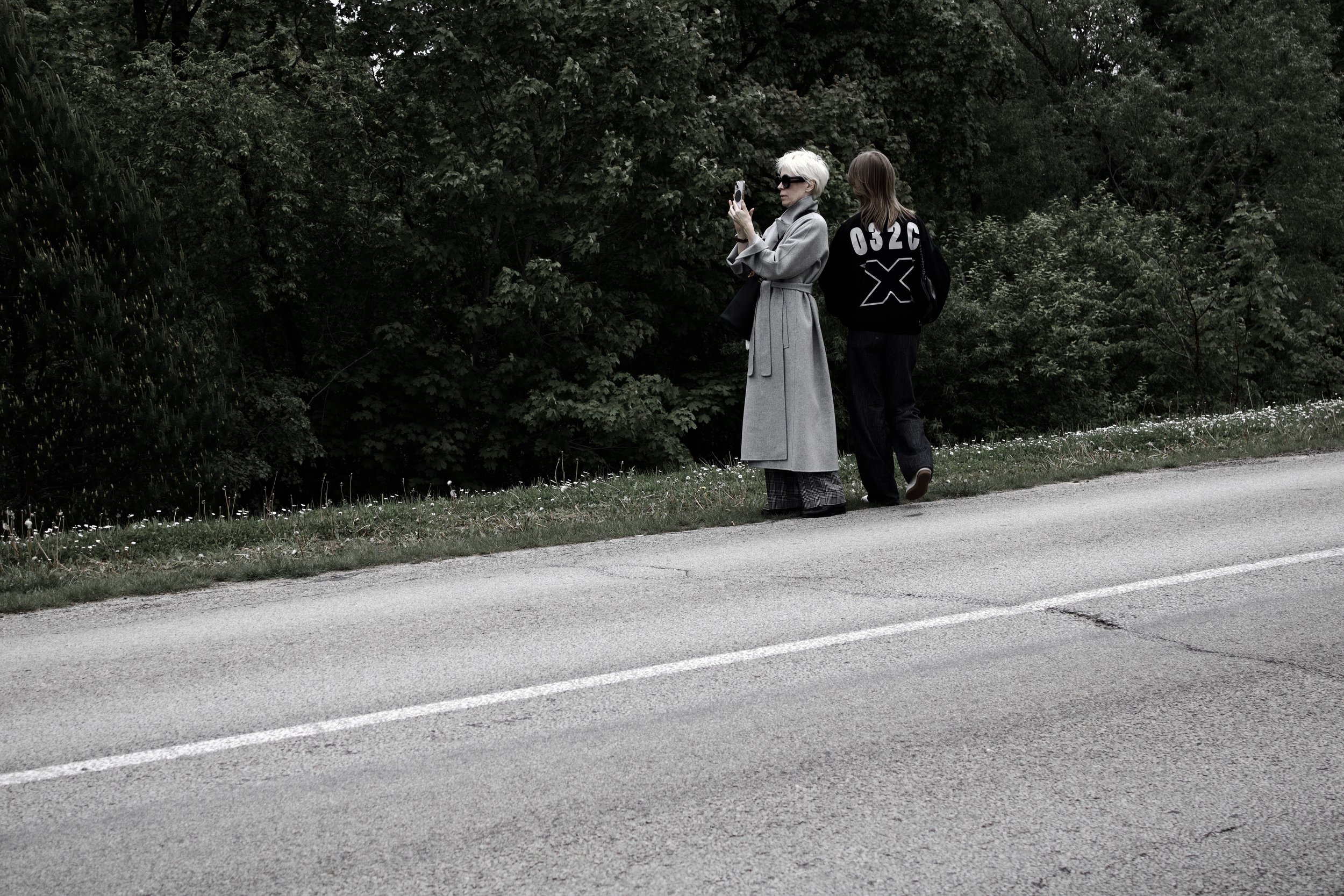Two women standing on the side of a road near dense greenery. One woman with short white hair is taking a photo with her phone, wearing a long gray coat and sunglasses. The other woman has long brown hair and is wearing a black jacket with white desi