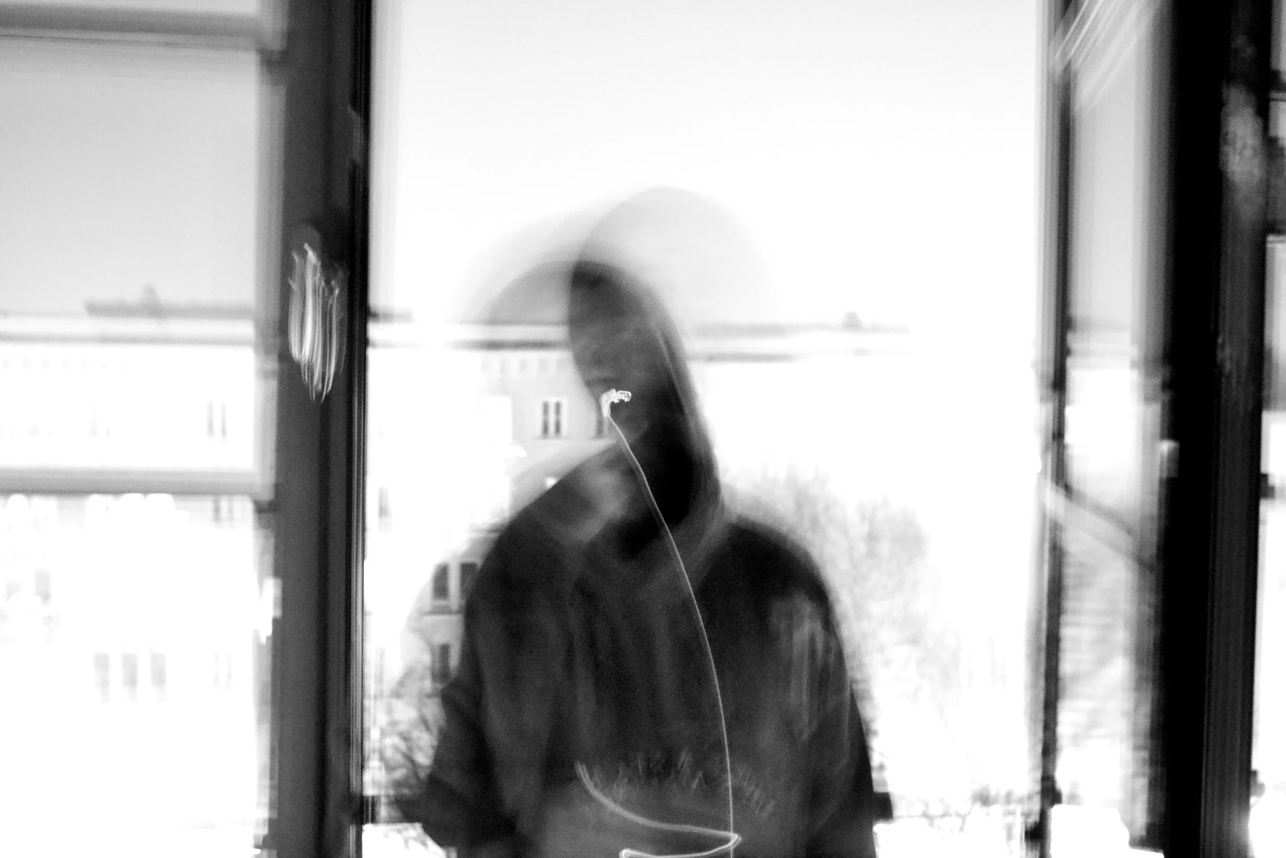 Black and white photo of a person taking a selfie in front of a window, with buildings visible outside.