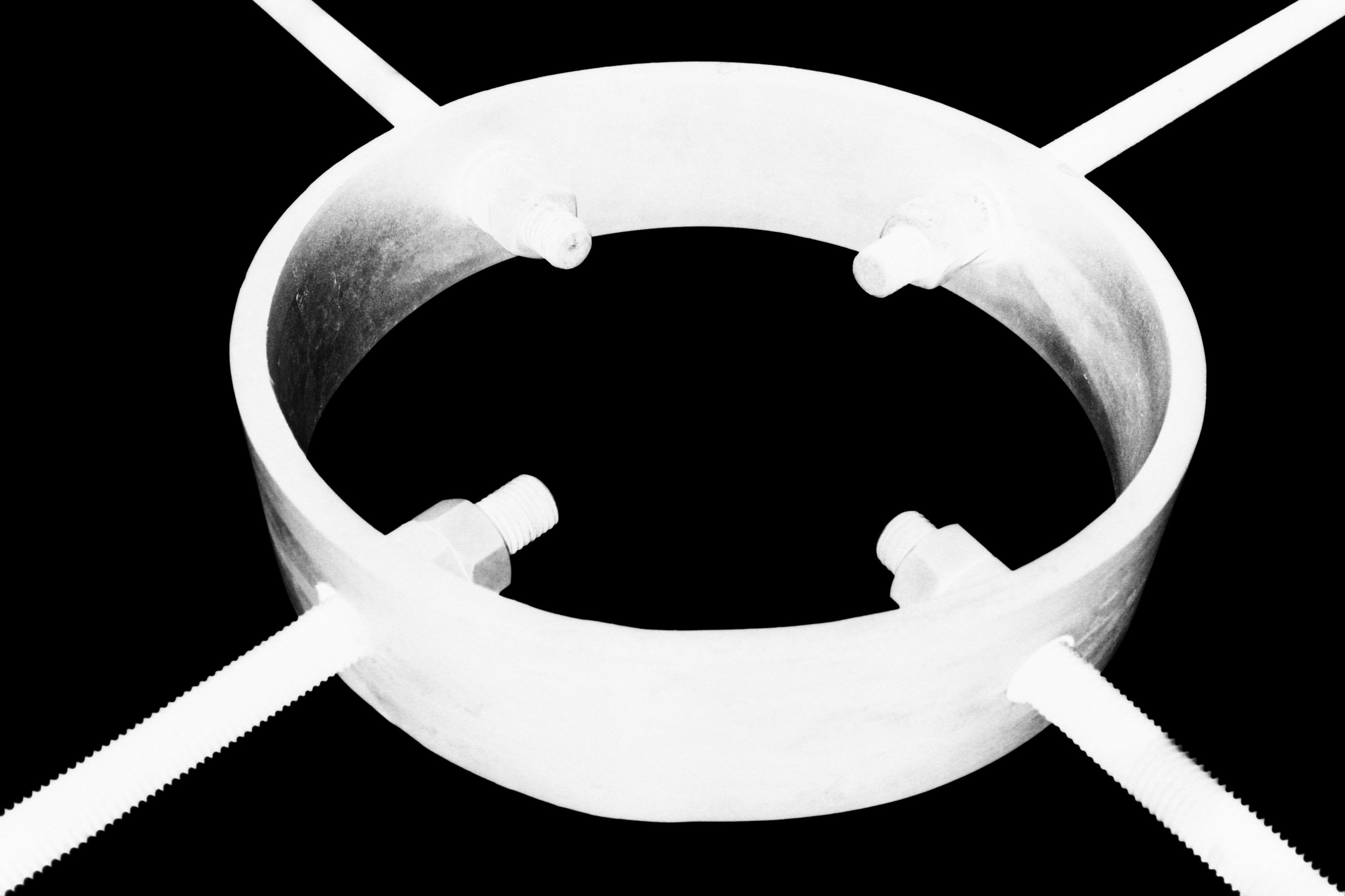 A close-up of a metallic ring with four bolts attached to it. Each bolt is secured through the ring and is extending outward. The image is inverted, black and white.
