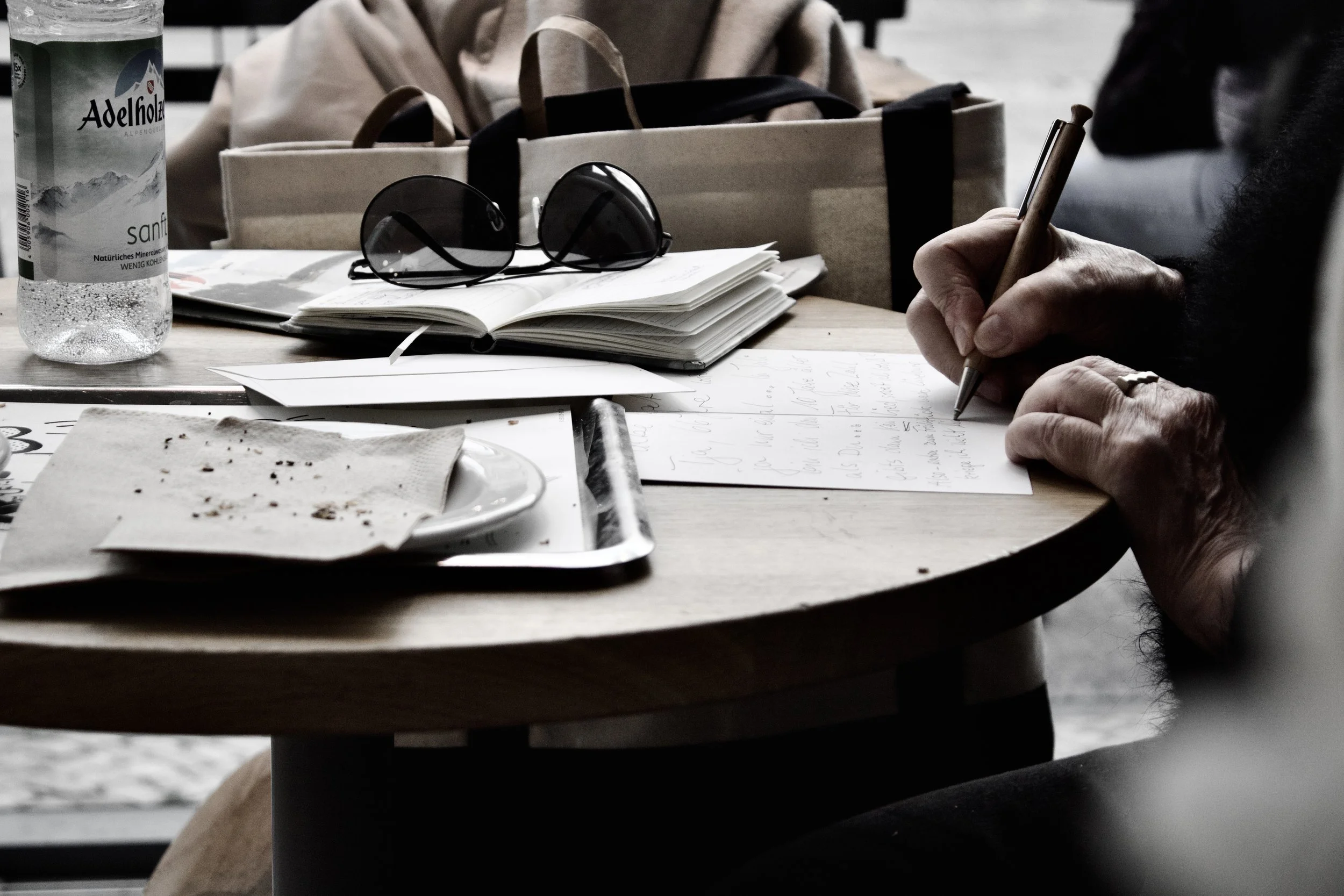 A person writes on a piece of paper at a round table, with sunglasses, a bottle of water, and open notebooks nearby.
